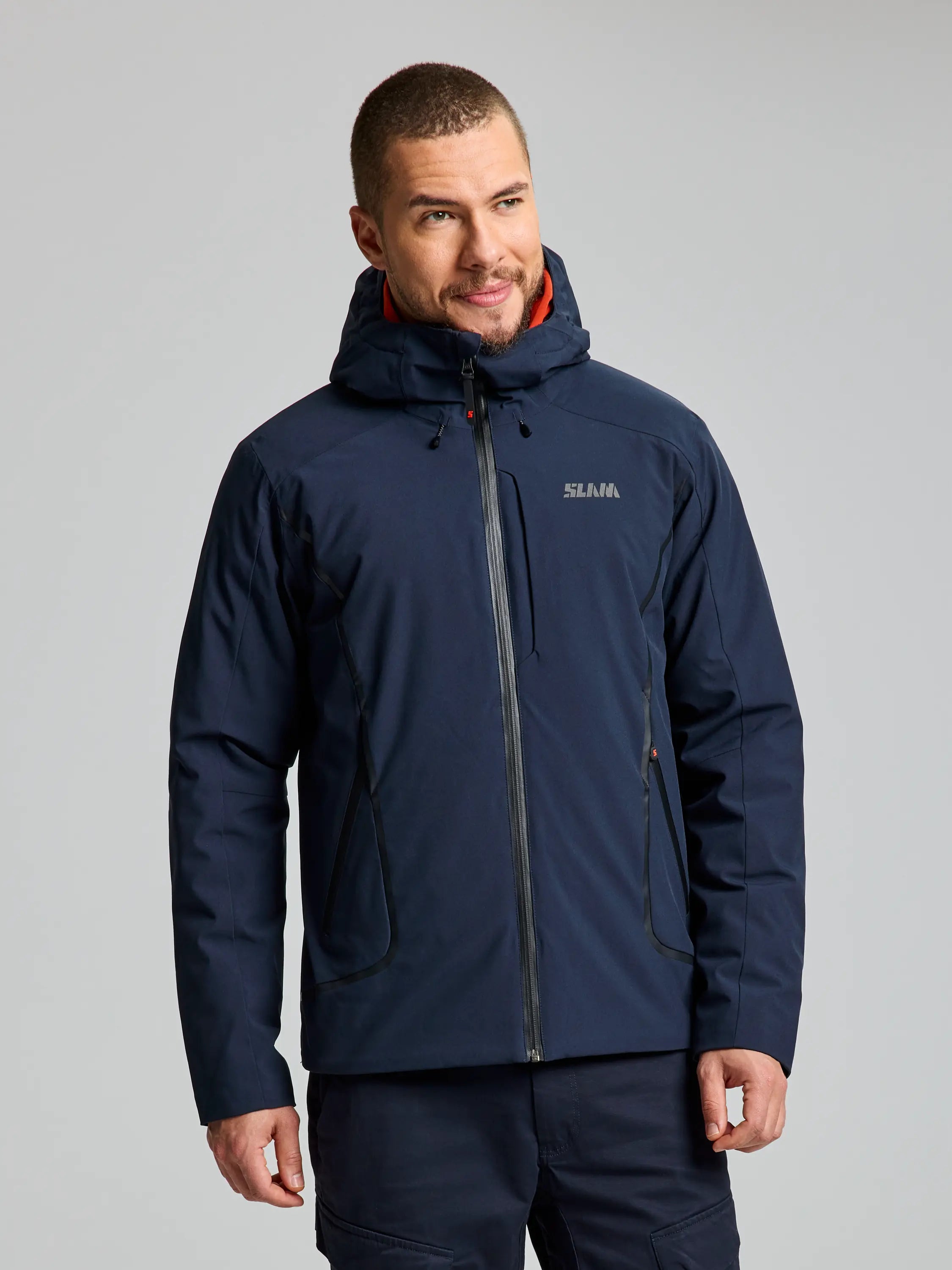 A person wearing a DARK NAVY ACTIVE HOODED INSULATED JACKET by SLAM stands against a plain background. This waterproof jacket features a zipper, two pockets, and a logo on the chest. The individual, with short hair and a trimmed beard, exudes style while enjoying the breathability of their attire.