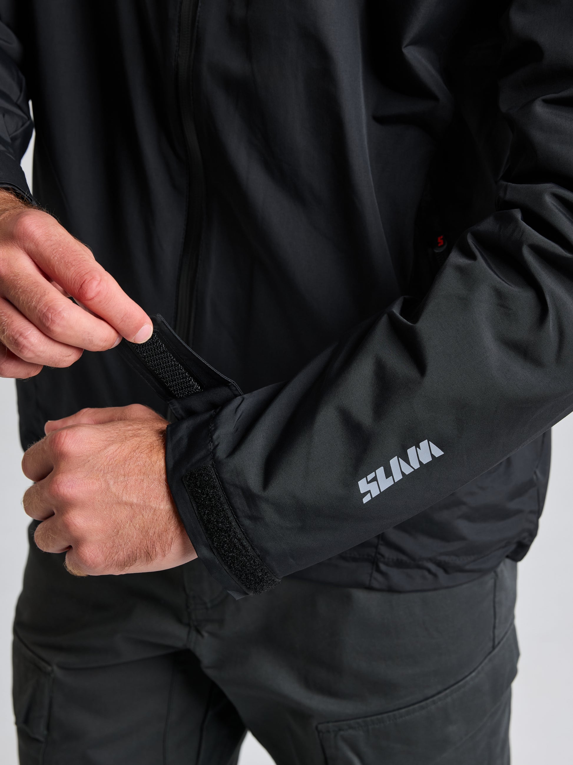 A person adjusts the Velcro cuff of a black CREW WARM HOODED JACKET, featuring the brand name "SLAM" on the sleeve. The hands and part of the torso are visible against a plain background.