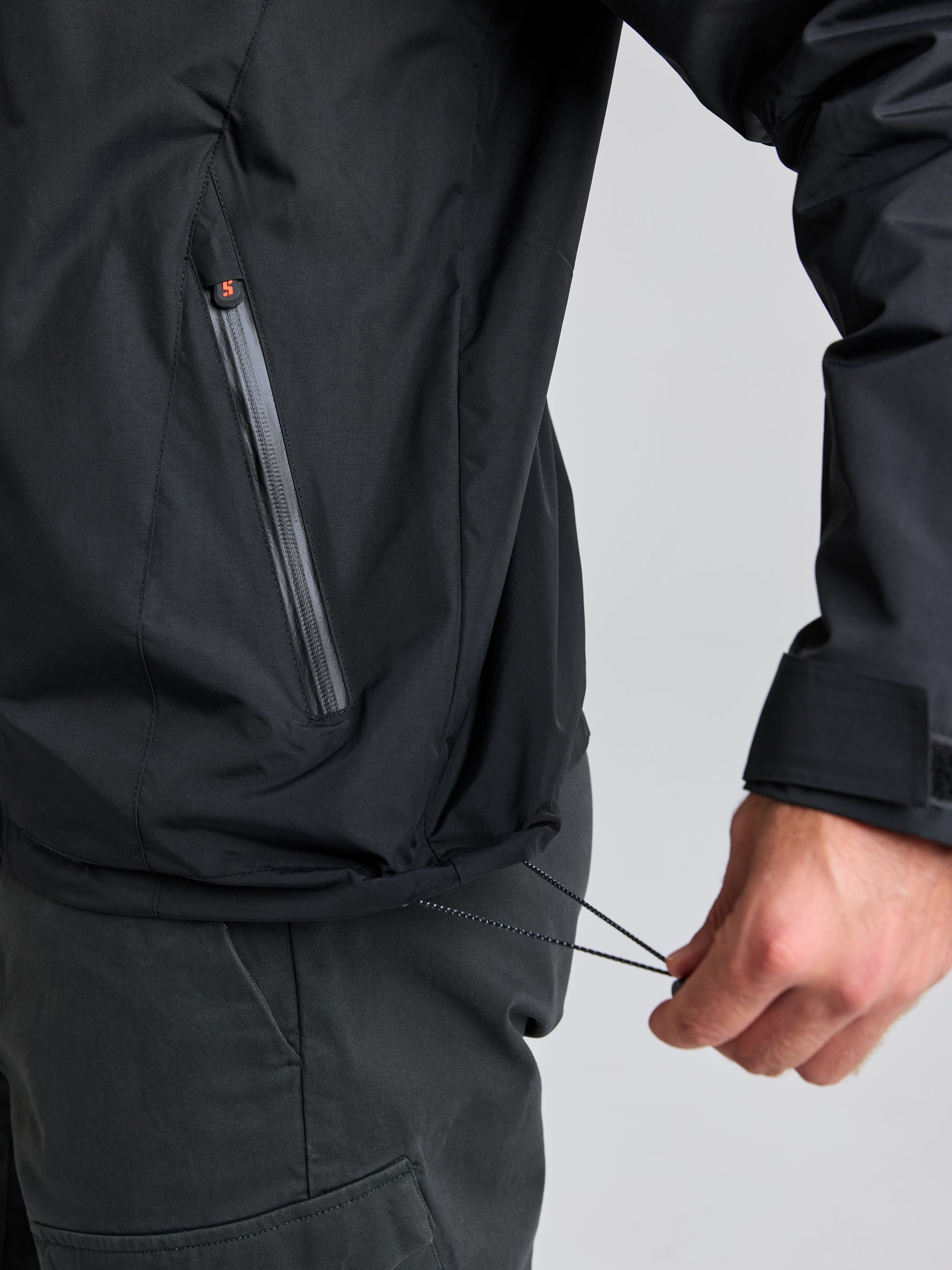 A person adjusts the drawstring on the hem of a SLAM CREW WARM HOODED JACKET, perfect for brisk days out at sea. The black winter sailing jacket features a zippered pocket on the side and pairs seamlessly with their dark pants against the plain background.