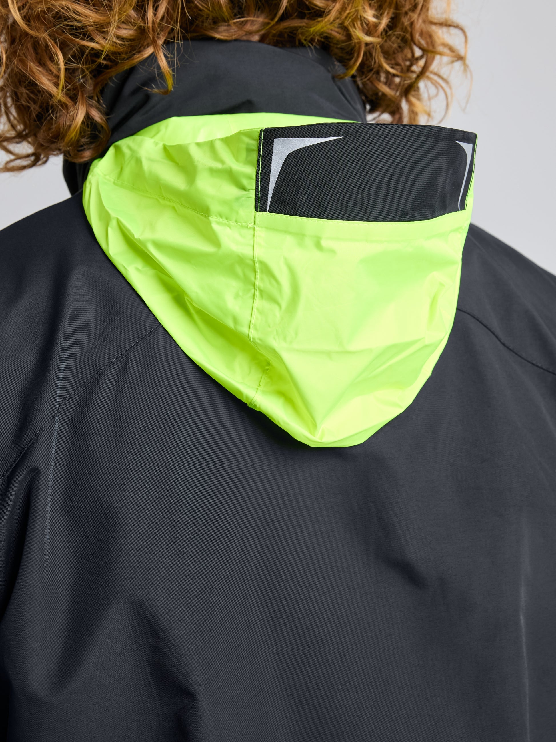 A close-up shows a person donning the SLAM CREW WARM HOODED JACKET, featuring a black design with a vivid yellow-green hood. The back of the hood is adorned with a distinctive black and white pattern. Curly hair peeks out from the top, suggesting warm comfort beneath its waterproof surface.