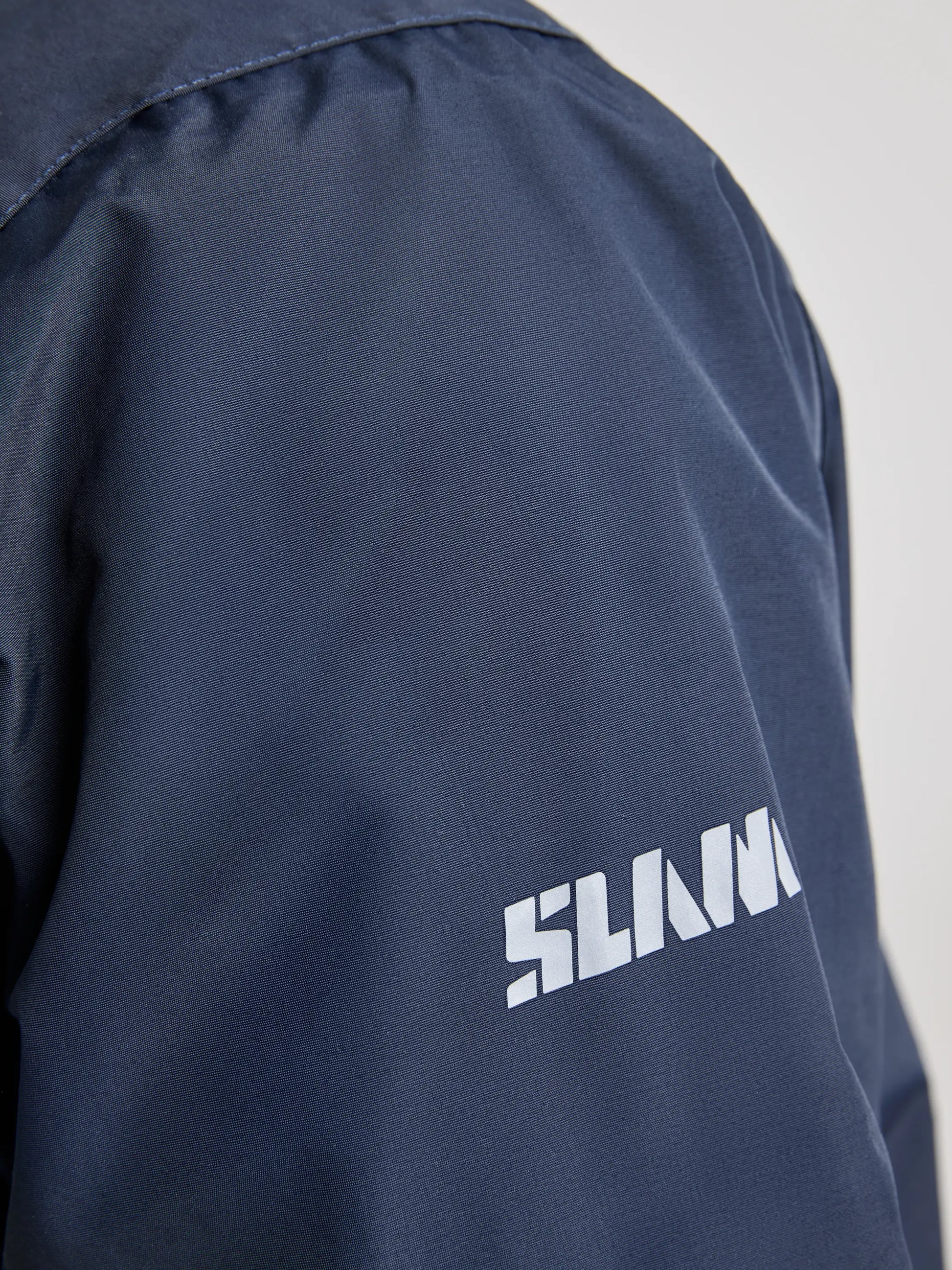 Close-up of a navy blue CREW WARM HOODED JACKET by SLAM, featuring white block letters spelling "SLAM" on the sleeve, expertly crafted from waterproof material for enhanced protection.