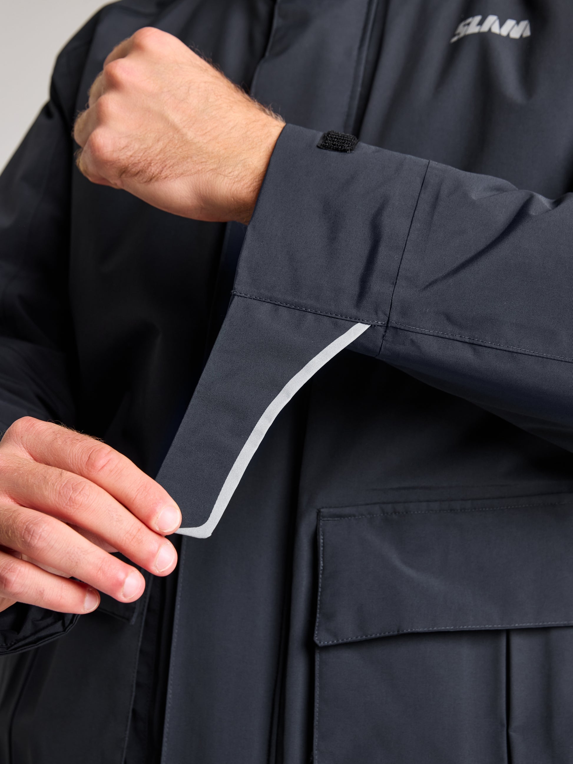 A person adjusts the sleeve of the SLAM ACT ETNZ LONG PARKA, a navy blue waterproof jacket, pulling on the velcro strap. This jacket provides breathable comfort and features visible stitching along with a pocket on its lower part. Only the hands and part of the arms are visible.