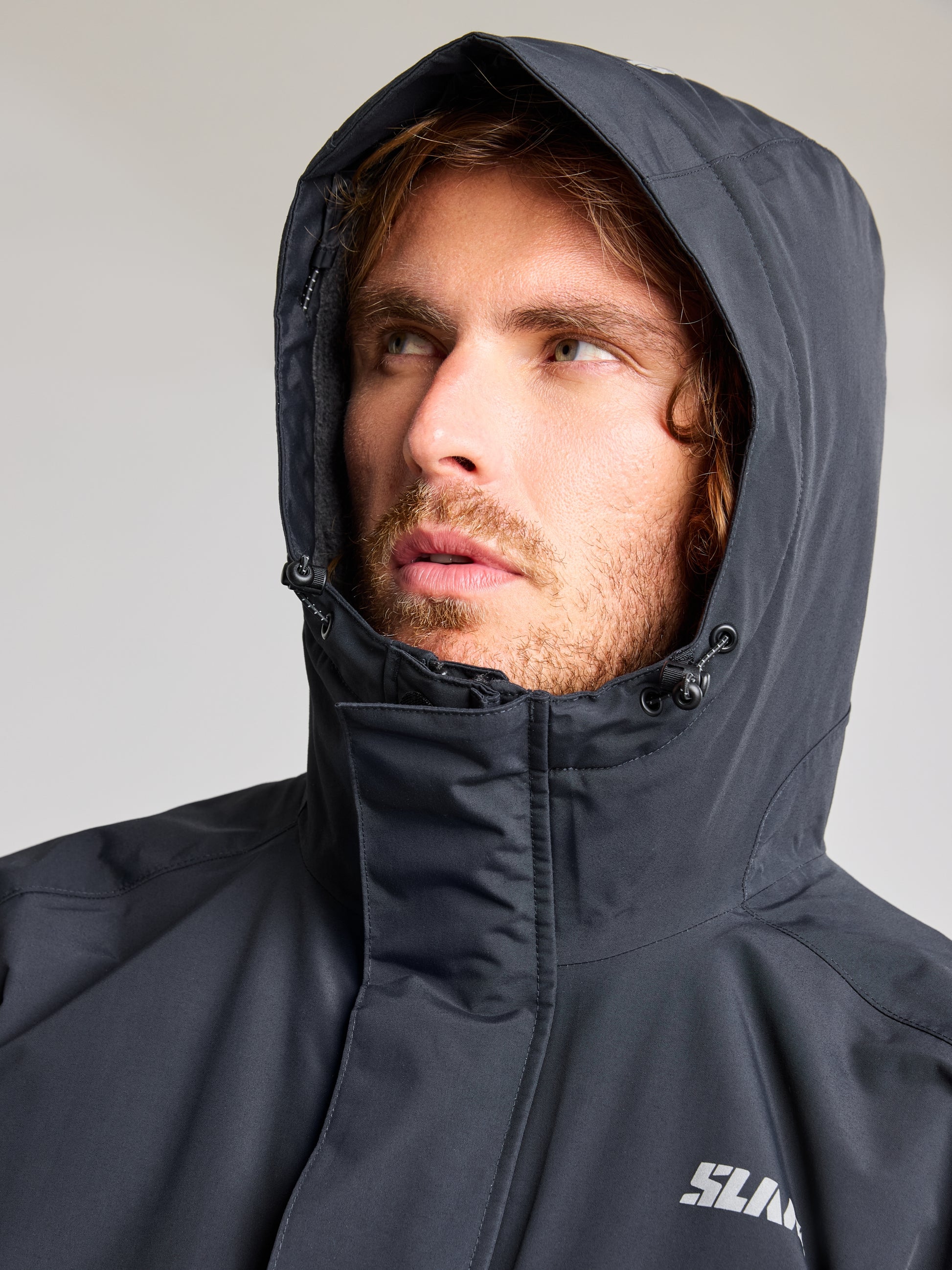 A person with long hair is wearing an ACT ETNZ LONG PARKA by SLAM, looking slightly to the side. The breathable parka features a zipper and drawstring, with a small logo on the left chest. The background is plain and neutral.