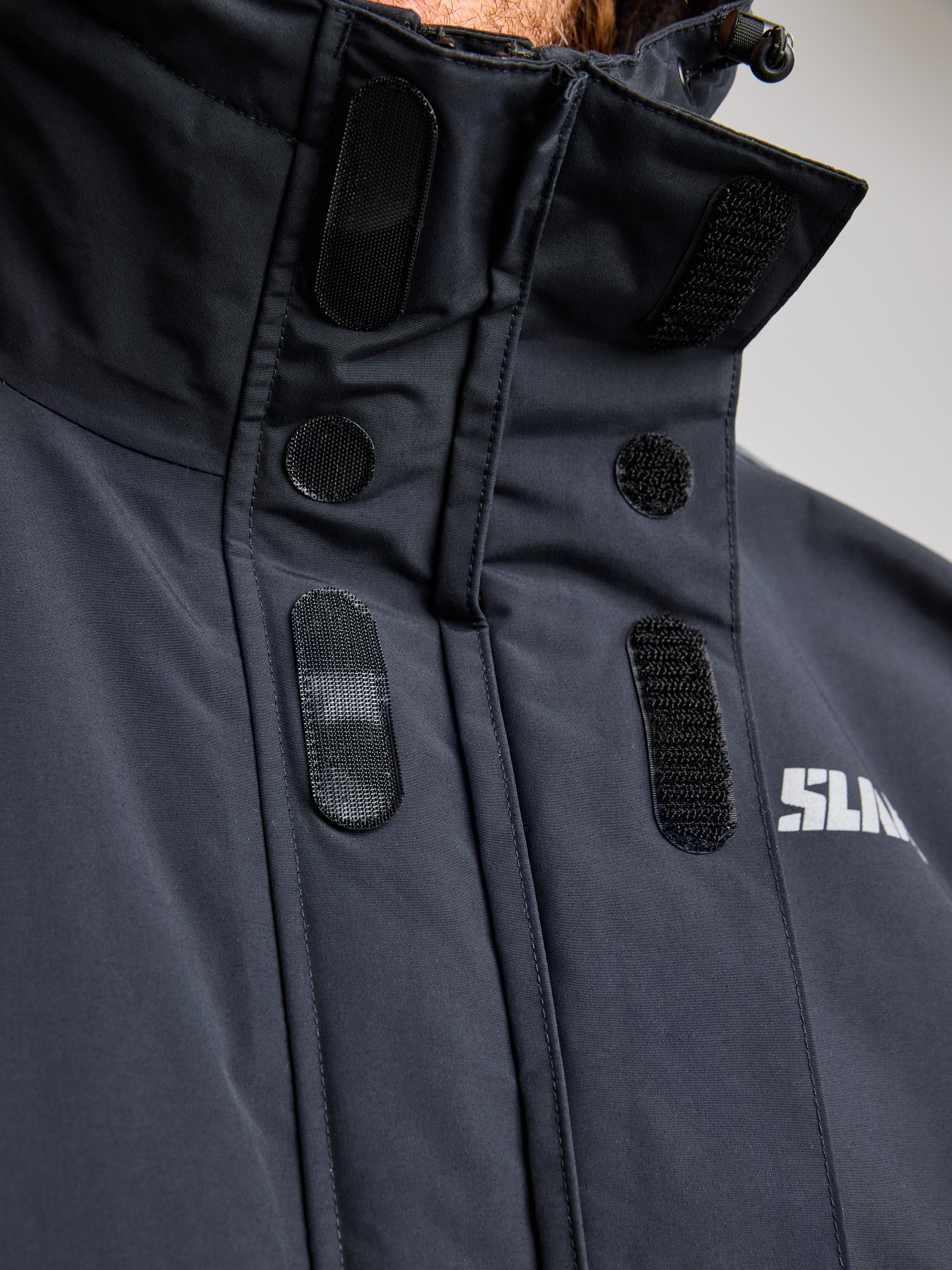 Close-up of the ACT ETNZ LONG PARKA in black by SLAM, featuring Velcro fastenings on the collar designed to keep you warm and dry. The high-neck style highlights its breathable fabric, with a partial view of a printed white logo on the left side, concentrating on texture and fastening details.