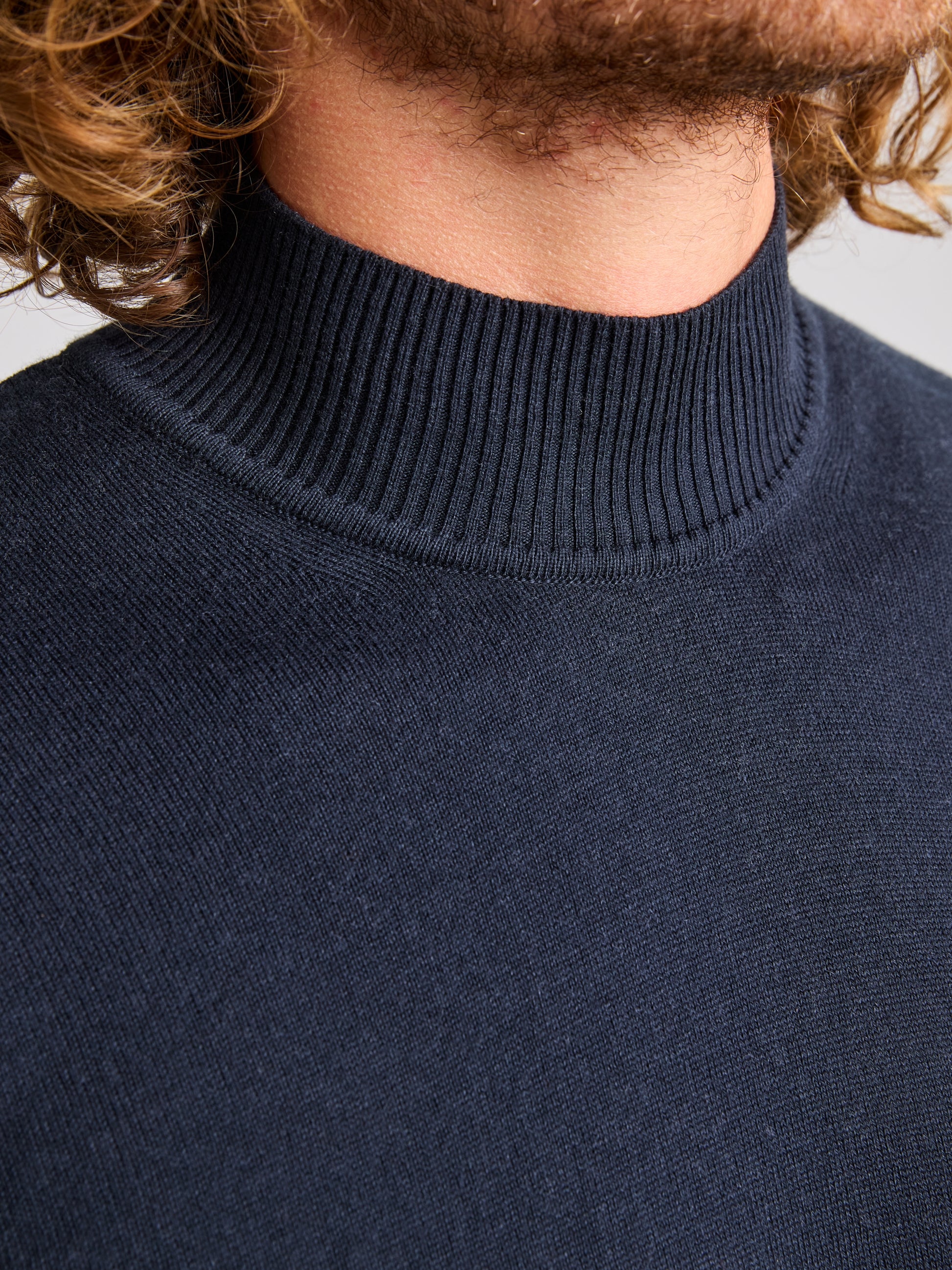 Close-up of a person wearing the JUMPER ERSE CL by SLAM in navy blue. The focus is on the ribbed collar, with the person's curly hair partially visible above the neckline. This classic men's design stands out against a neutral, softly blurred background.