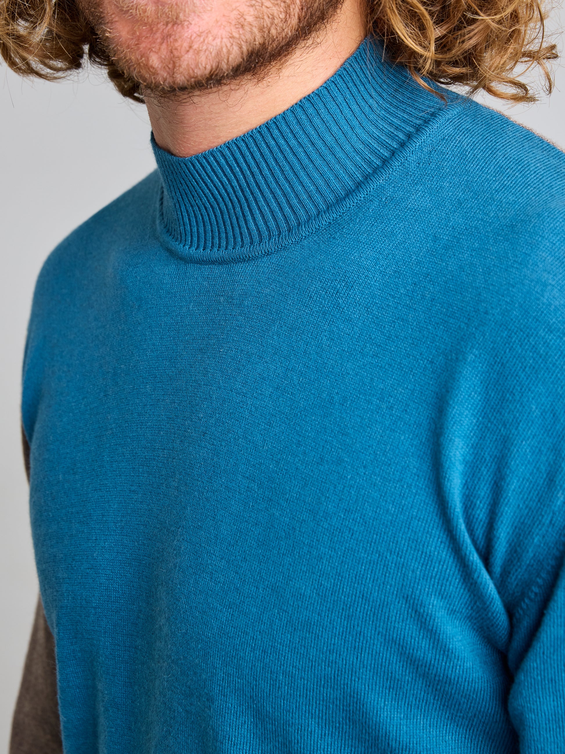 A person with wavy, light brown hair is wearing a teal JUMPER ERSE CL from the SLAM brand. The image is cropped to show only part of their face and upper torso, emphasizing the stylish design of the men's sweater. The background is a plain gray.