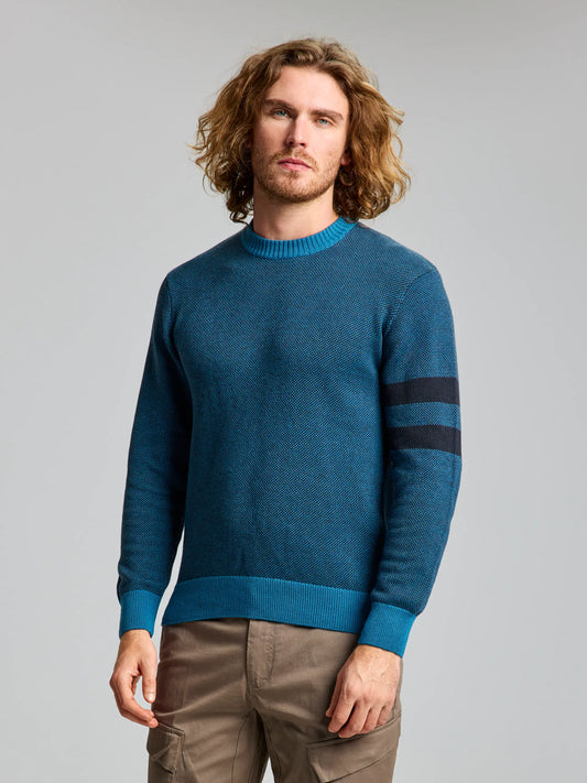 A person with shoulder-length curly hair is wearing the JUMPER RUDRA CL by SLAM, a Polynesia-colored sweater crafted from a cotton/wool blend with dark horizontal stripes on one sleeve. They are also dressed in khaki pants, set against a plain gray background.