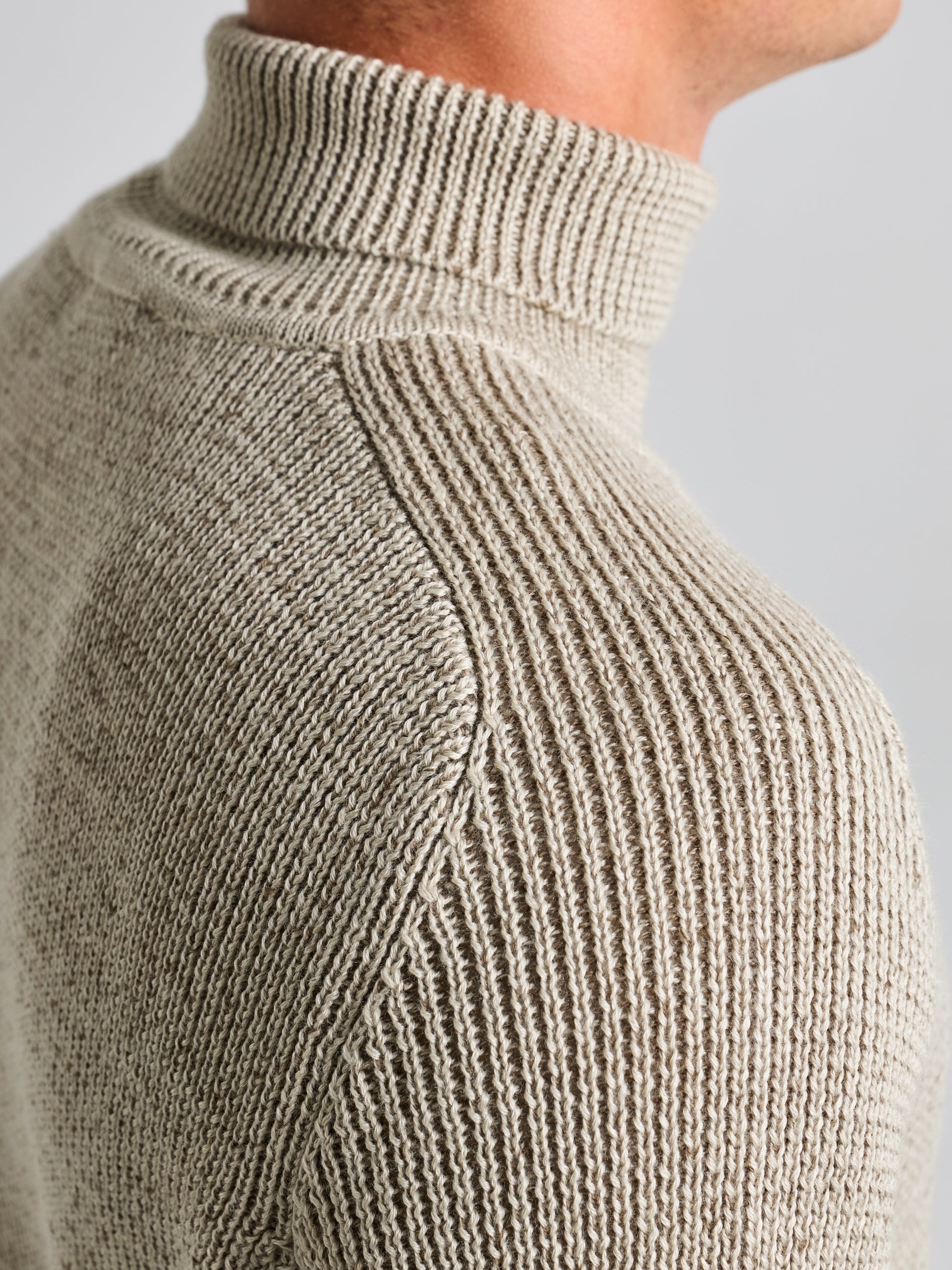 A close-up features a person wearing a beige SEAWOOL TURTLENECK by SLAM, highlighting the intricate ribbed texture of the Merino blend yarn, with a focus on the shoulder seam and neckline against a neutral light gray background.