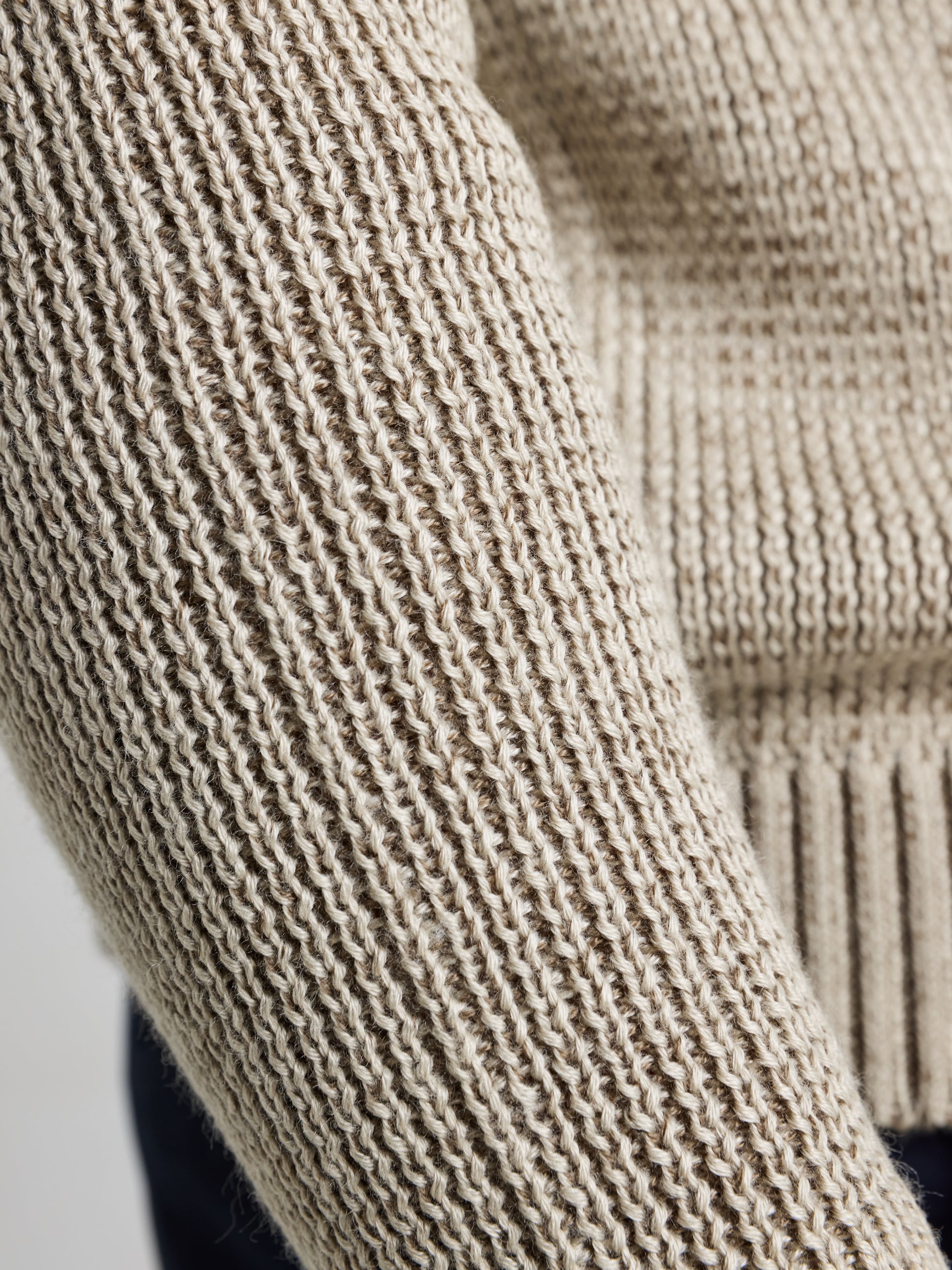 A close-up image showcases the detailed texture of beige, ribbed knit fabric from SLAM's SEAWOOL TURTLENECK, highlighting individual knit stitches and a section of the cuff. Crafted with recycled Seawool, this piece offers both warmth and sustainability.