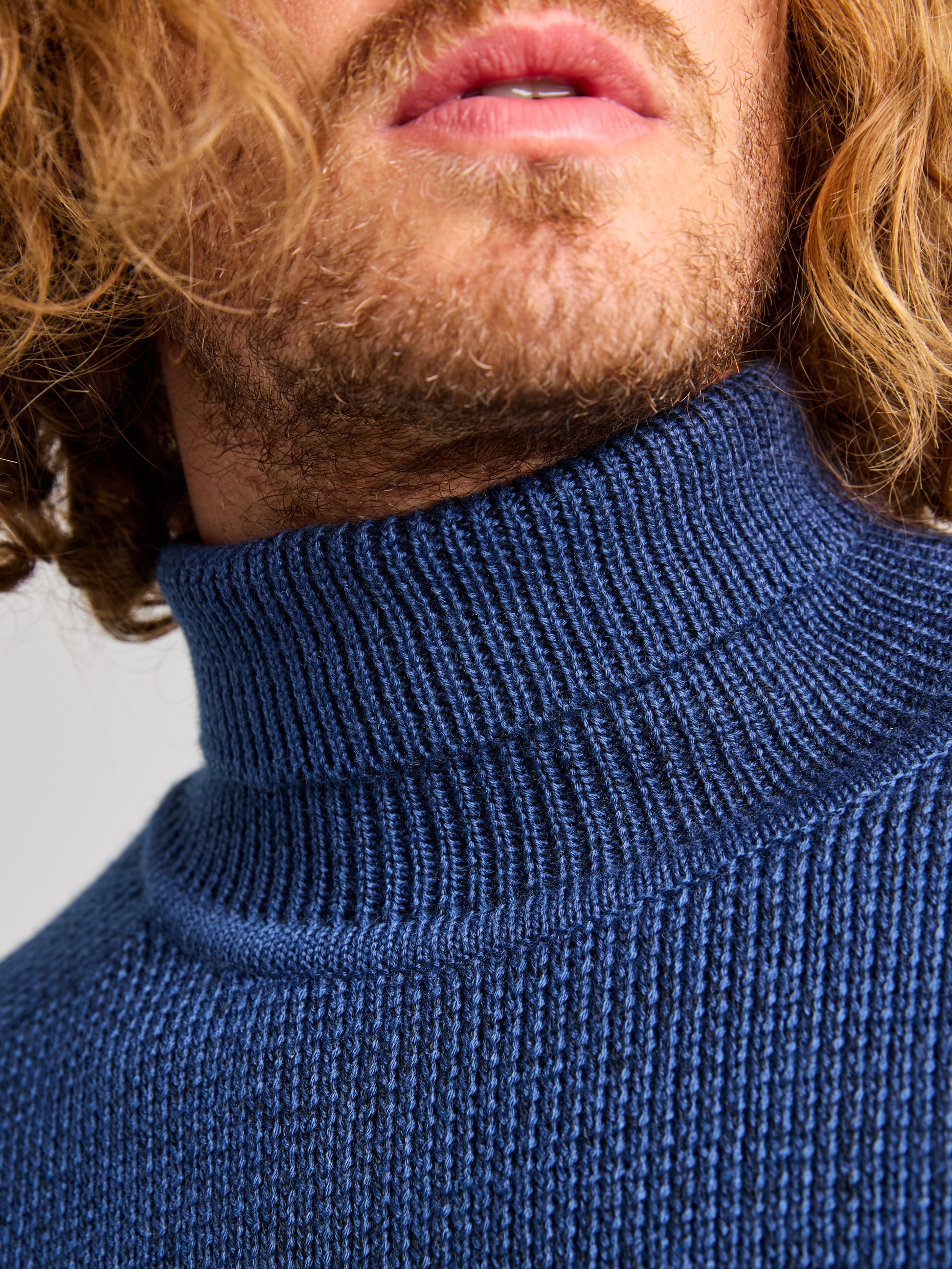 Close-up of a person's lower face with a light brown mustache and beard, wearing a SLAM SEAWOOL TURTLENECK crafted from recycled Seawool. Their shoulder-length, light brown hair is wavy.