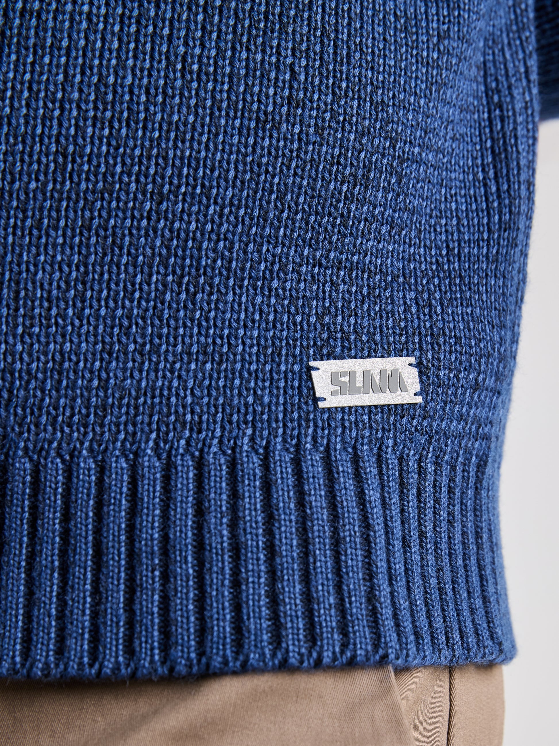 A close-up of the SEAWOOL TURTLENECK by SLAM reveals its blue hue made from recycled Seawool, complete with a ribbed hem and a small white logo label sewn near it. Designed for style and sustainability, it is paired with light brown pants.