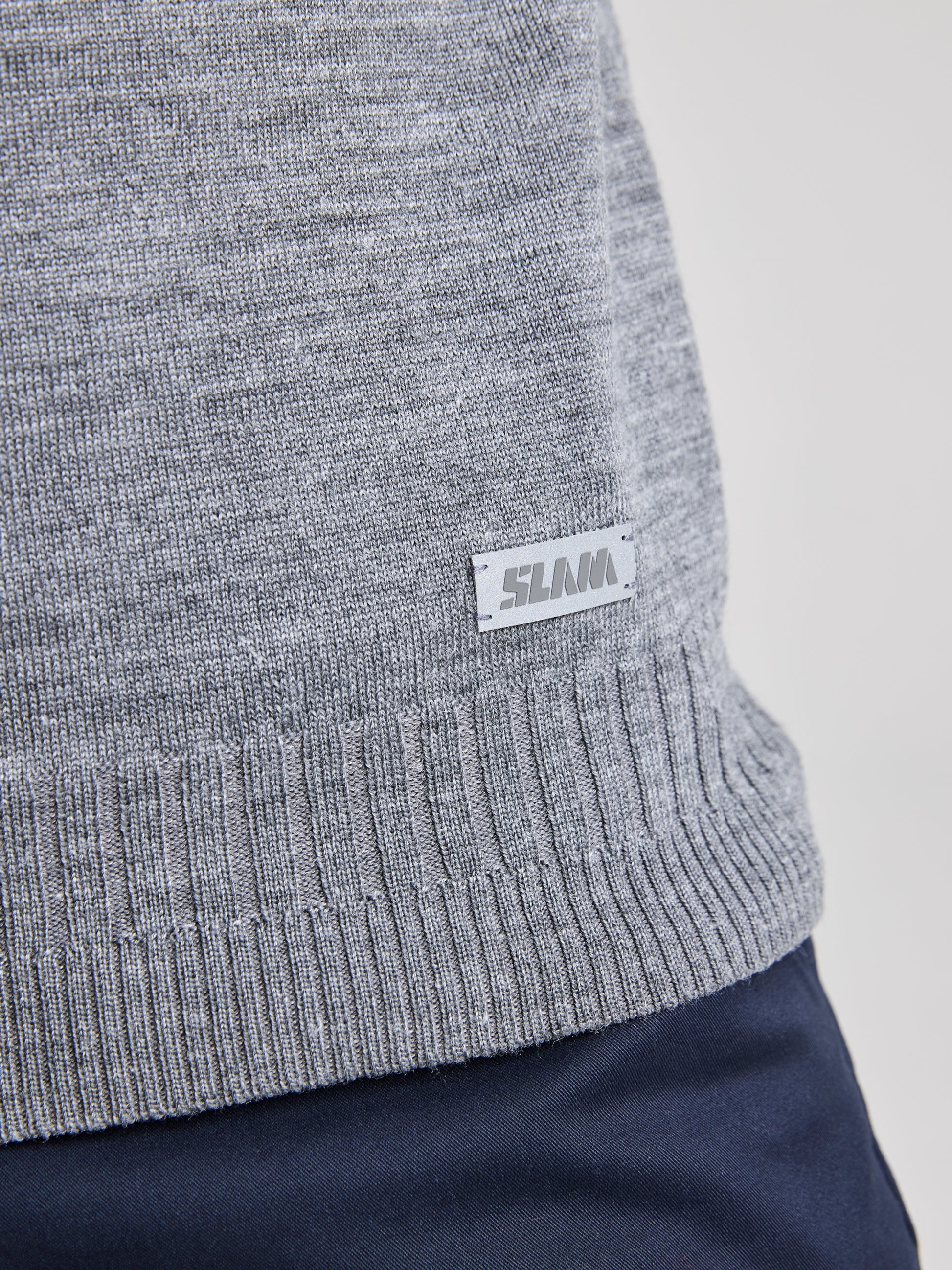 Close-up of an individual wearing the SLAM SEAWOOL CREWNECK sweater in gray, made from Merino-blend yarn with ribbed hem details and featuring a small "SLAM" tag on the side, paired with dark blue regular fit pants.