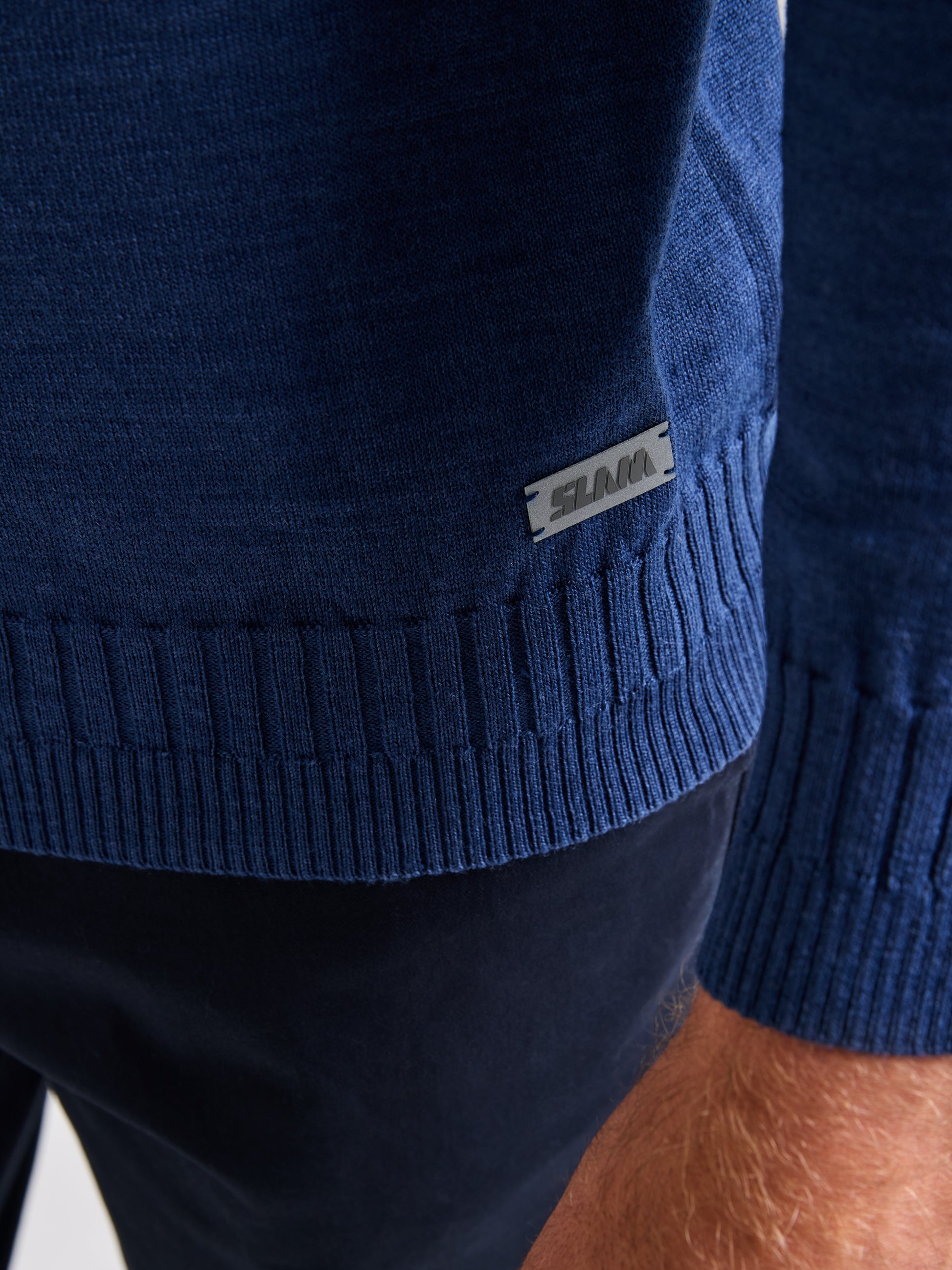 Close-up of a person wearing a dark blue SLAM SEAWOOL CREWNECK sweater and black pants. The hem, made from Merino-blend yarn, showcases ribbed detailing and includes a small metal tag with text on it. The person's hand is visible next to the sweater.