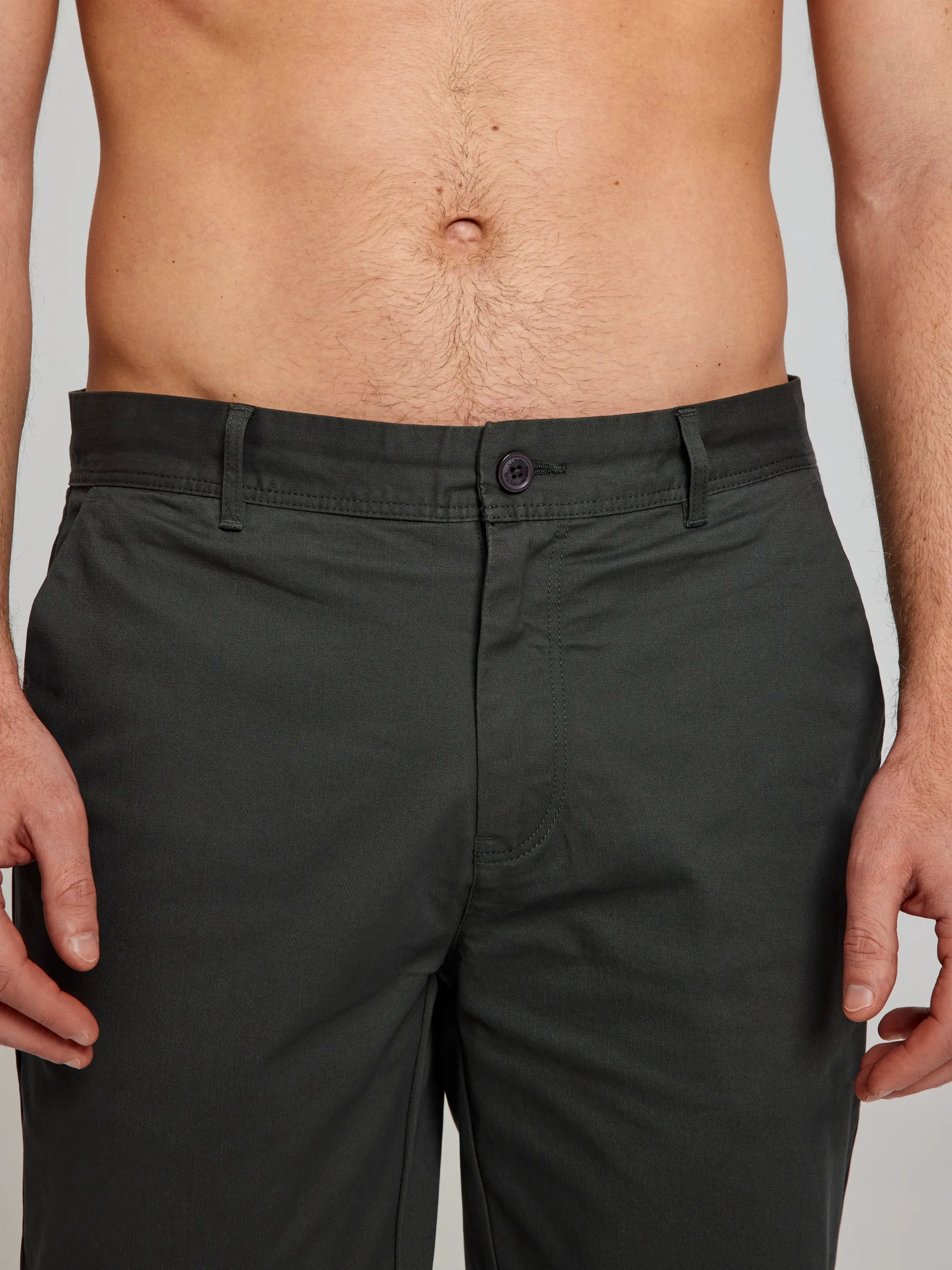 A person wearing SLAM's DECK CHINO PANT CL in dark green cotton demonstrates a comfortable sporty fit. The emphasis is on the lower torso, with attention to the classic pants' button and zipper area. Their hands rest relaxed at their sides, while an unclothed upper body completes the casual look.