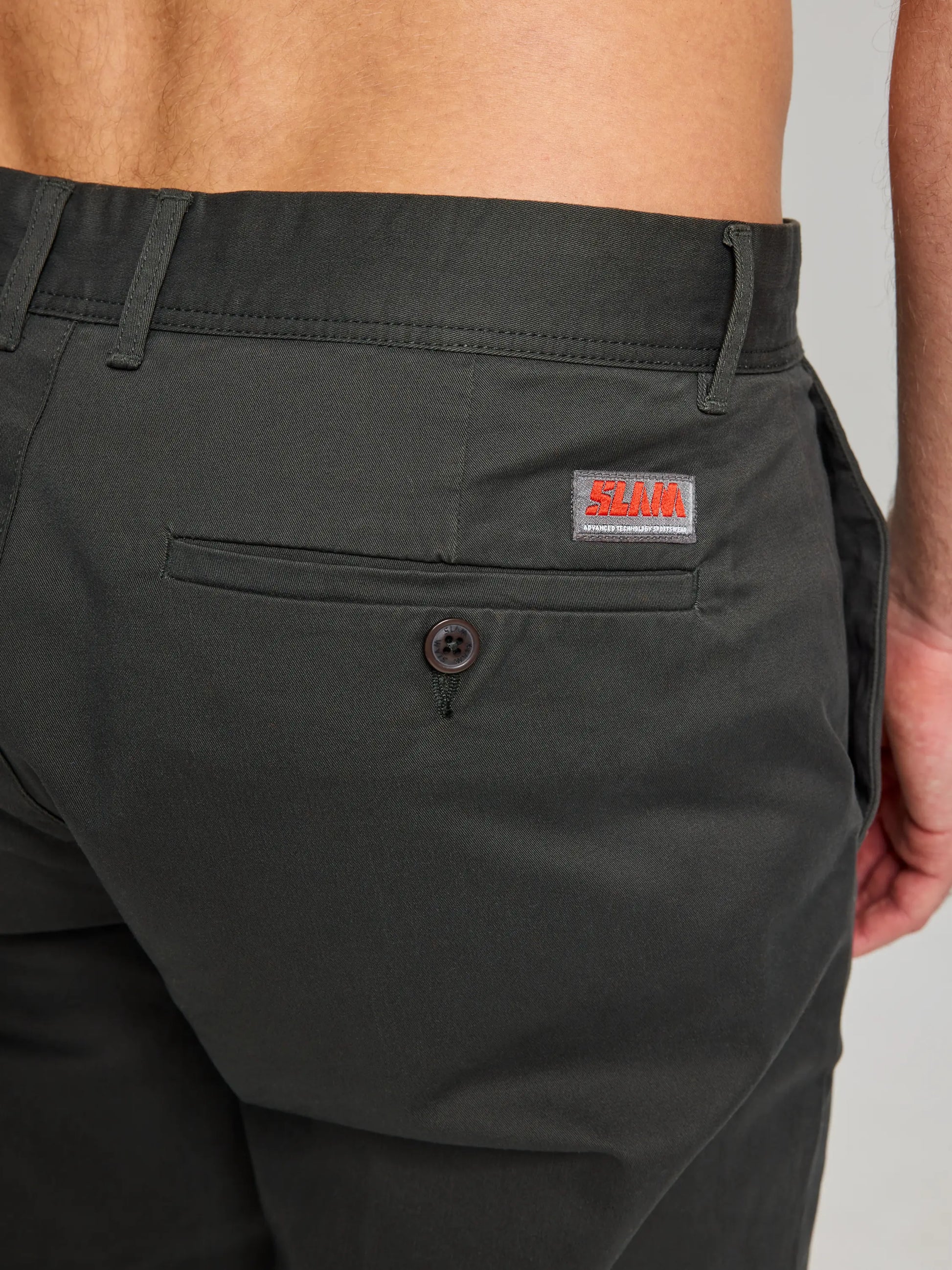 Close-up of a person wearing the DECK CHINO PANT CL in dark green, crafted from stretch cotton and designed with a waistband. These classic men's pants, from the SLAM brand, feature a back pocket with a button and an orange SLAM logo stitched above it. The person's back is partially visible, highlighting their comfortable sporty fit.