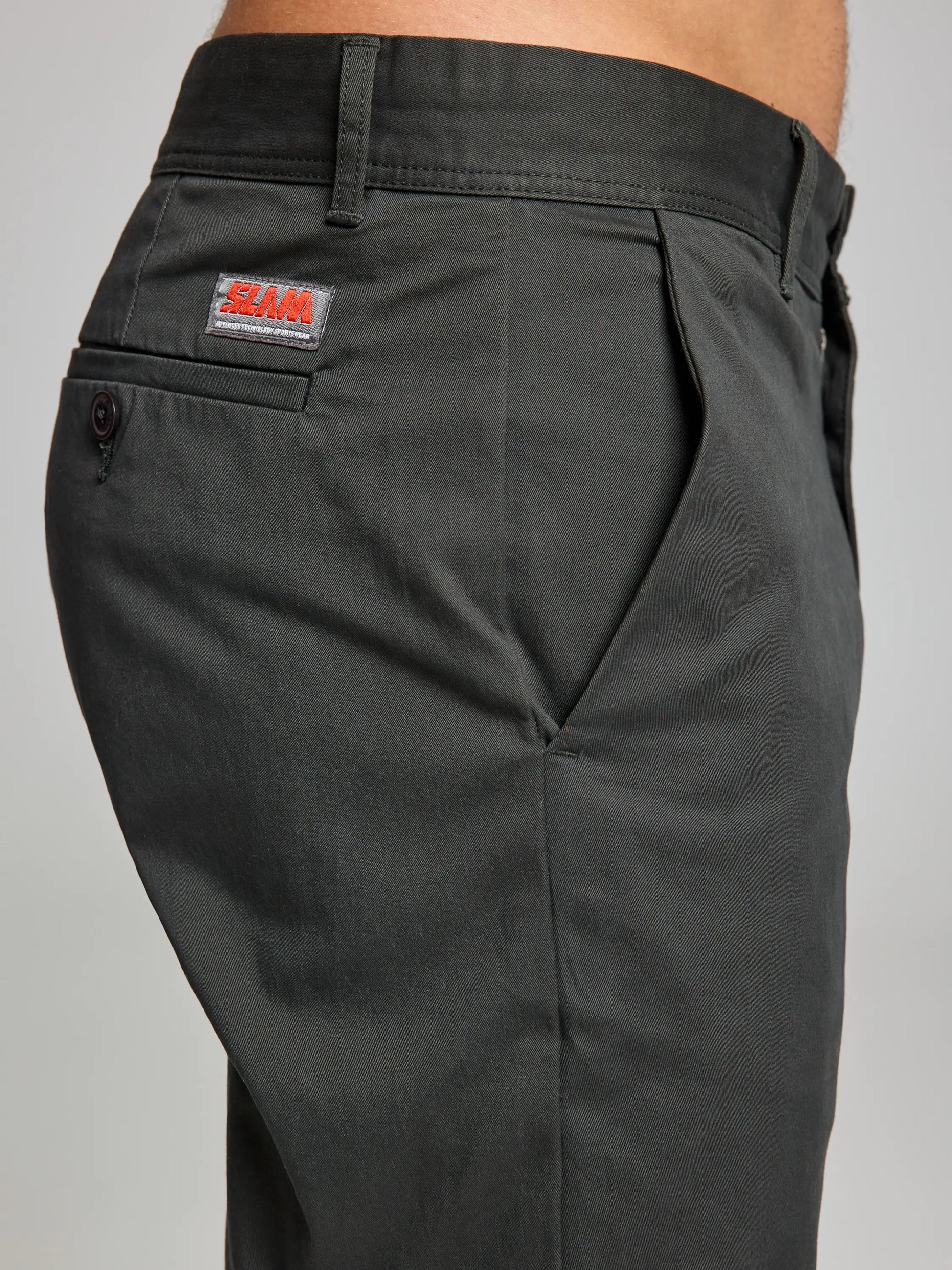 Side view of a person wearing the DECK CHINO PANT CL by SLAM in dark green, featuring a comfortable sporty fit. These trousers have a buttoned back pocket with a small red and white logo tag above it on the right side. Equipped with belt loops at the waistband, these stretch cotton chinos provide both style and comfort.