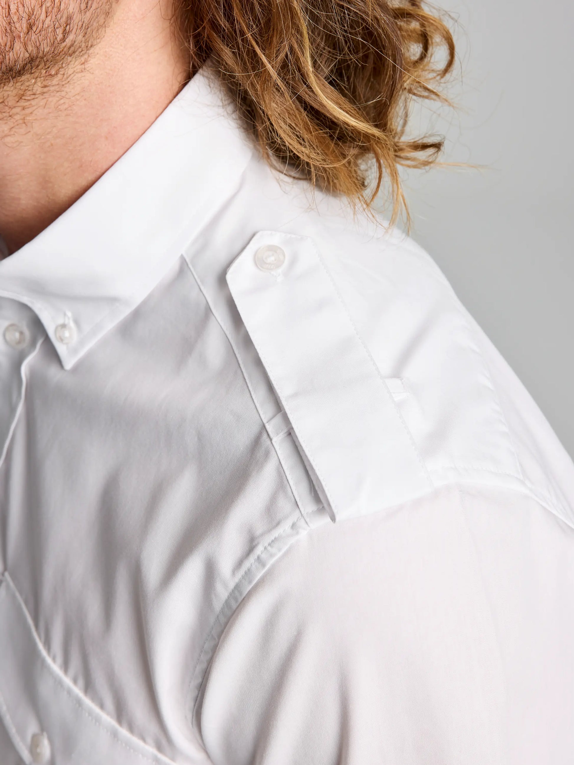 Close-up of a person wearing the SLAM DECK YACHT SHIRT CL, crafted in white cotton poplin with epaulets on the shoulders. The classic collar and visible stitching add detail, while long, wavy hair is partially visible. The background is plain and neutral, highlighting this stylish men's shirt.