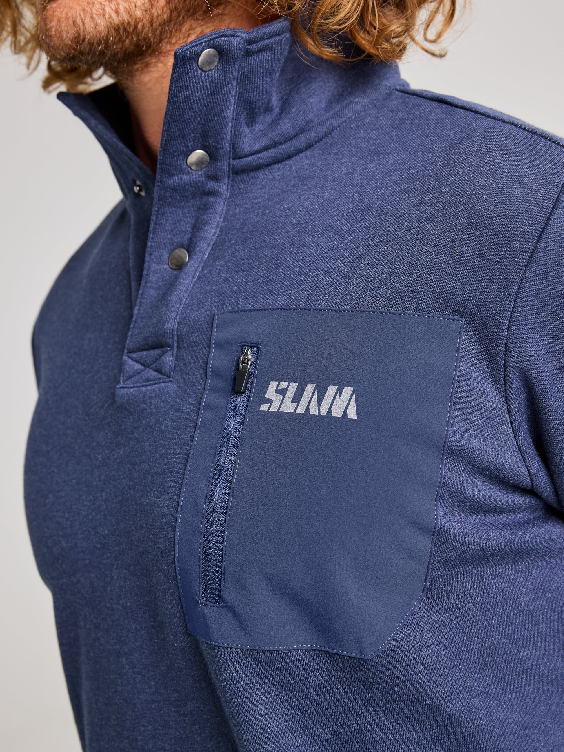 A person is wearing the SWEATSHIRT ANAPO CL, a blue men's sweatshirt from SLAM, featuring a high collar and 4-button opening. The garment includes a zippered chest pocket with "SLAM" printed on it. They have curly hair and are set against a plain gray background.