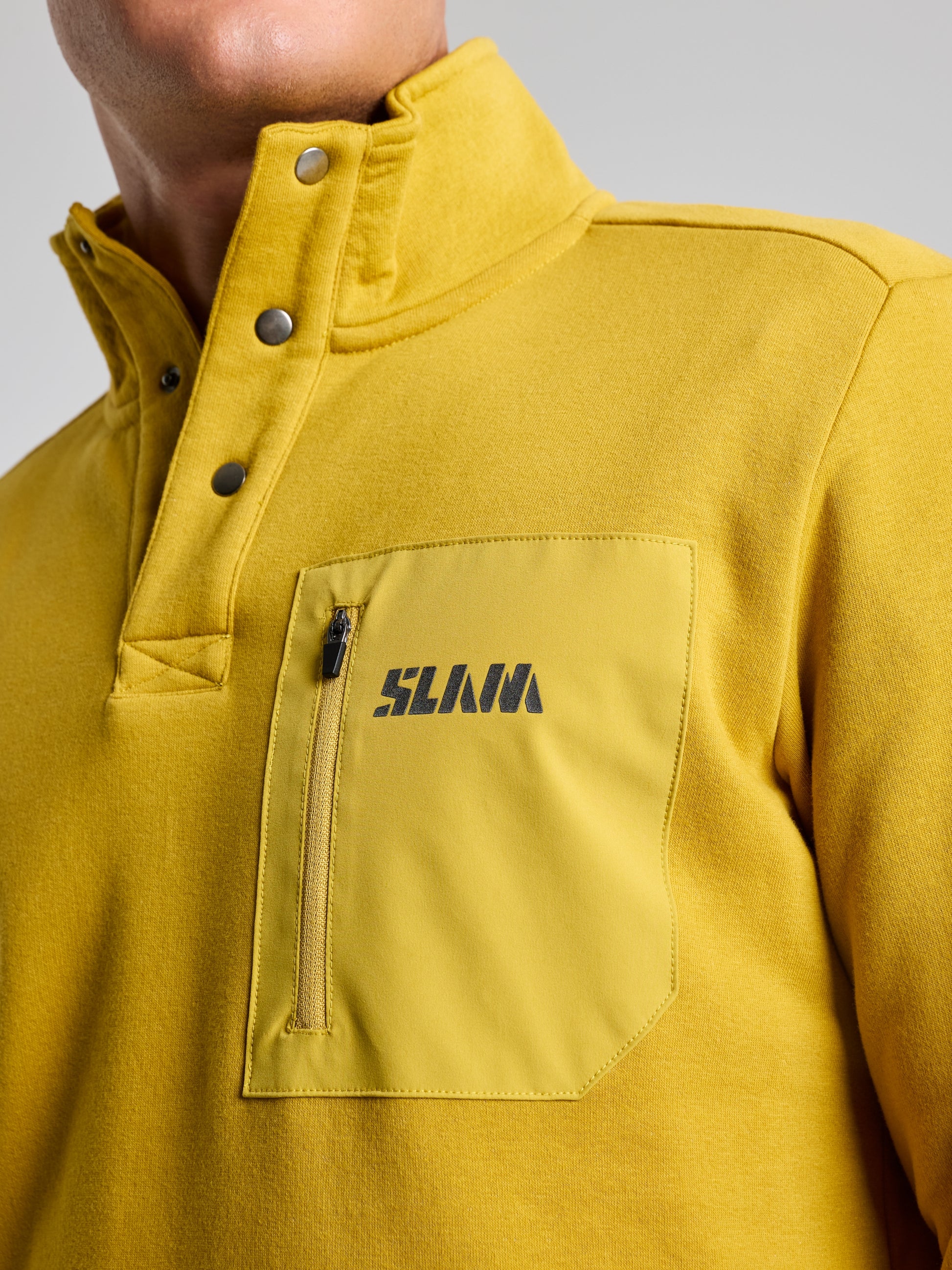 A person is wearing the SLAM brand's SWEATSHIRT ANAPO CL, which is a mustard yellow men's sweatshirt featuring a high collar and snap button opening. The garment includes a zippered chest pocket adorned with dark text, set against a neutral background.