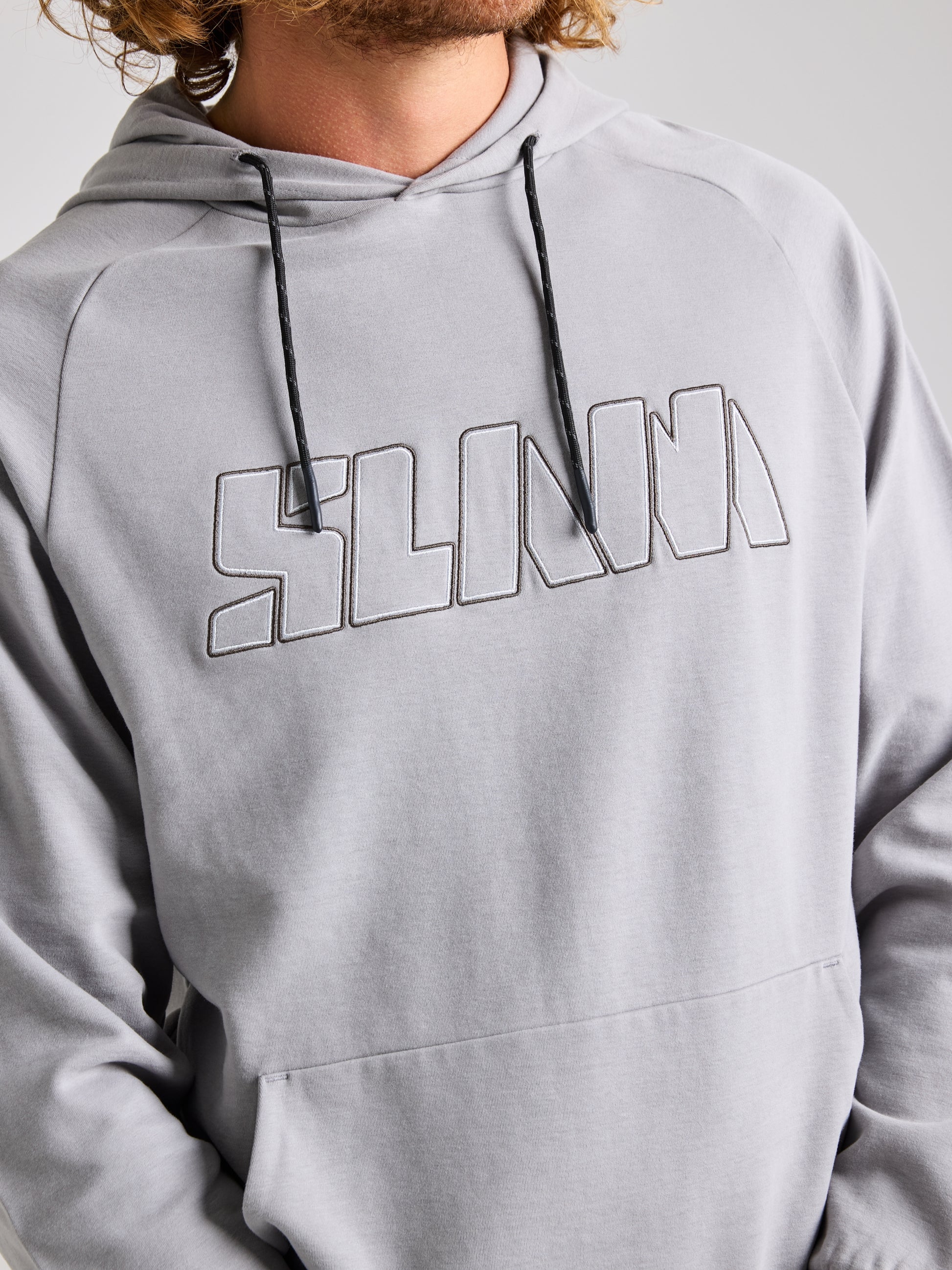 A person is dressed in a light gray men's LOGO HOODIE by SLAM, showcasing the word "SLINKY" prominently embroidered on the front in large capital letters. The hoodie is designed with an adjustable hood equipped with drawstrings, allowing their medium-length curly hair to peek through.