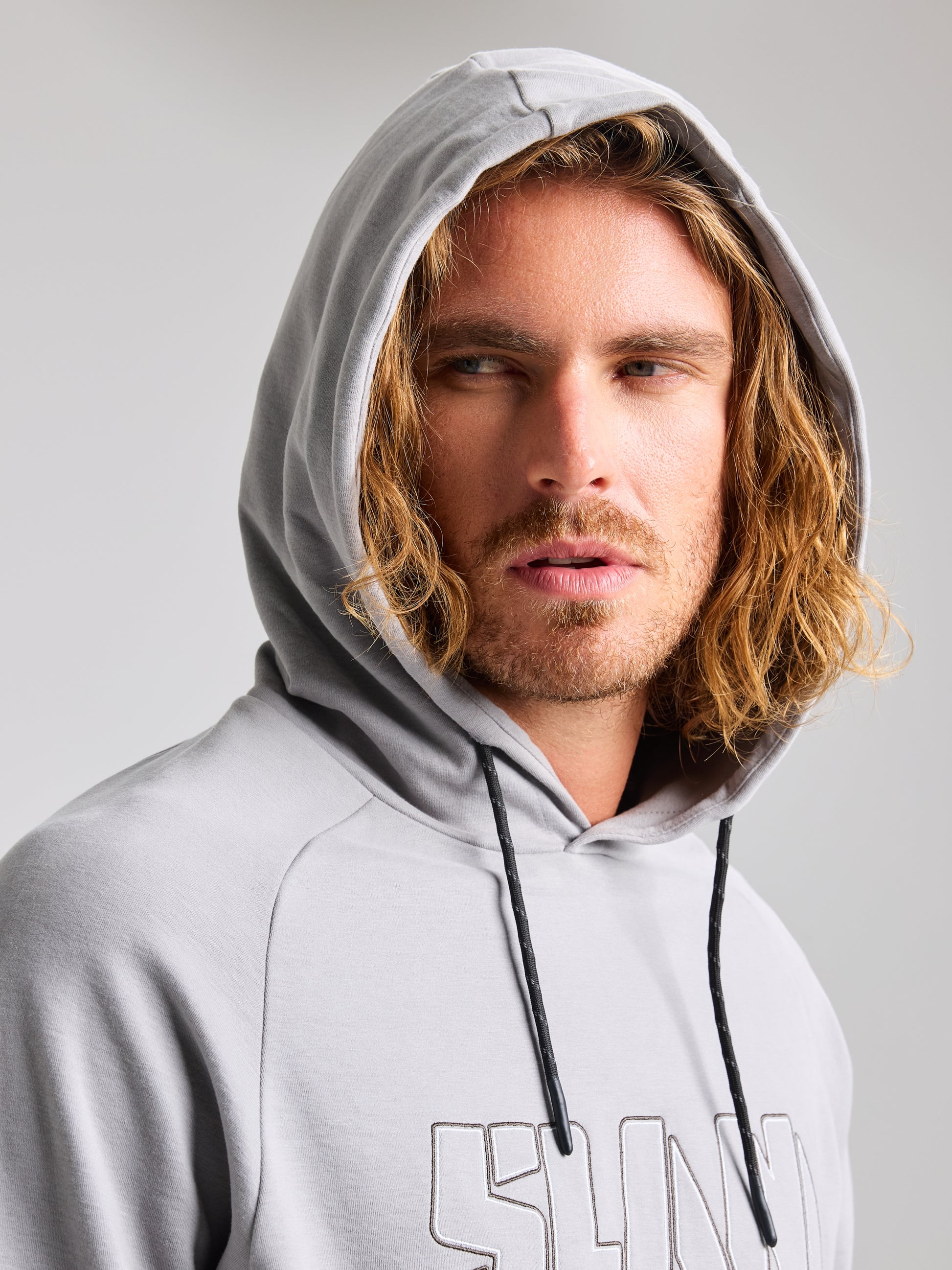 A person with long hair is wearing the SLAM LOGO HOODIE in gray, looking to the side against a neutral background. This men's warm sweatshirt features an adjustable hood for both comfort and style.