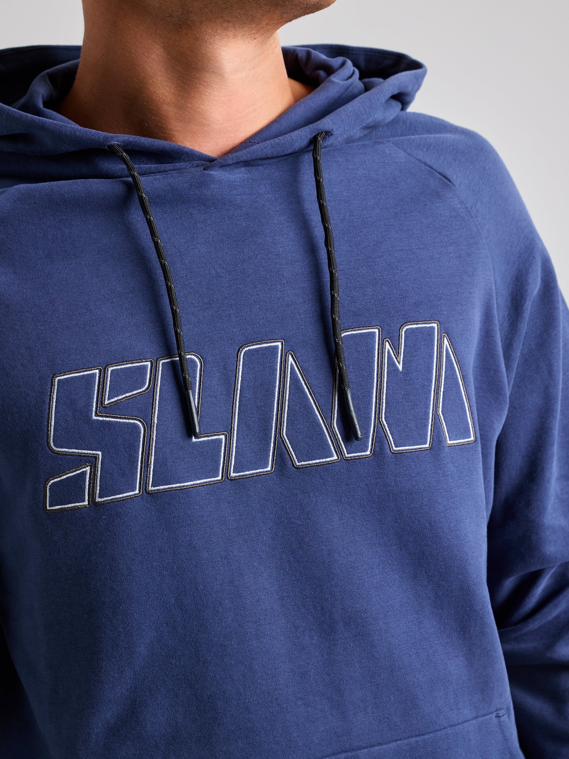 A person models the men's LOGO HOODIE by SLAM, which includes an adjustable hood and displays a geometric-style logo, with the bold word "SCALA" across the chest. The blue hoodie features a black drawstring and highlights only the lower portion of the person's face and neck.