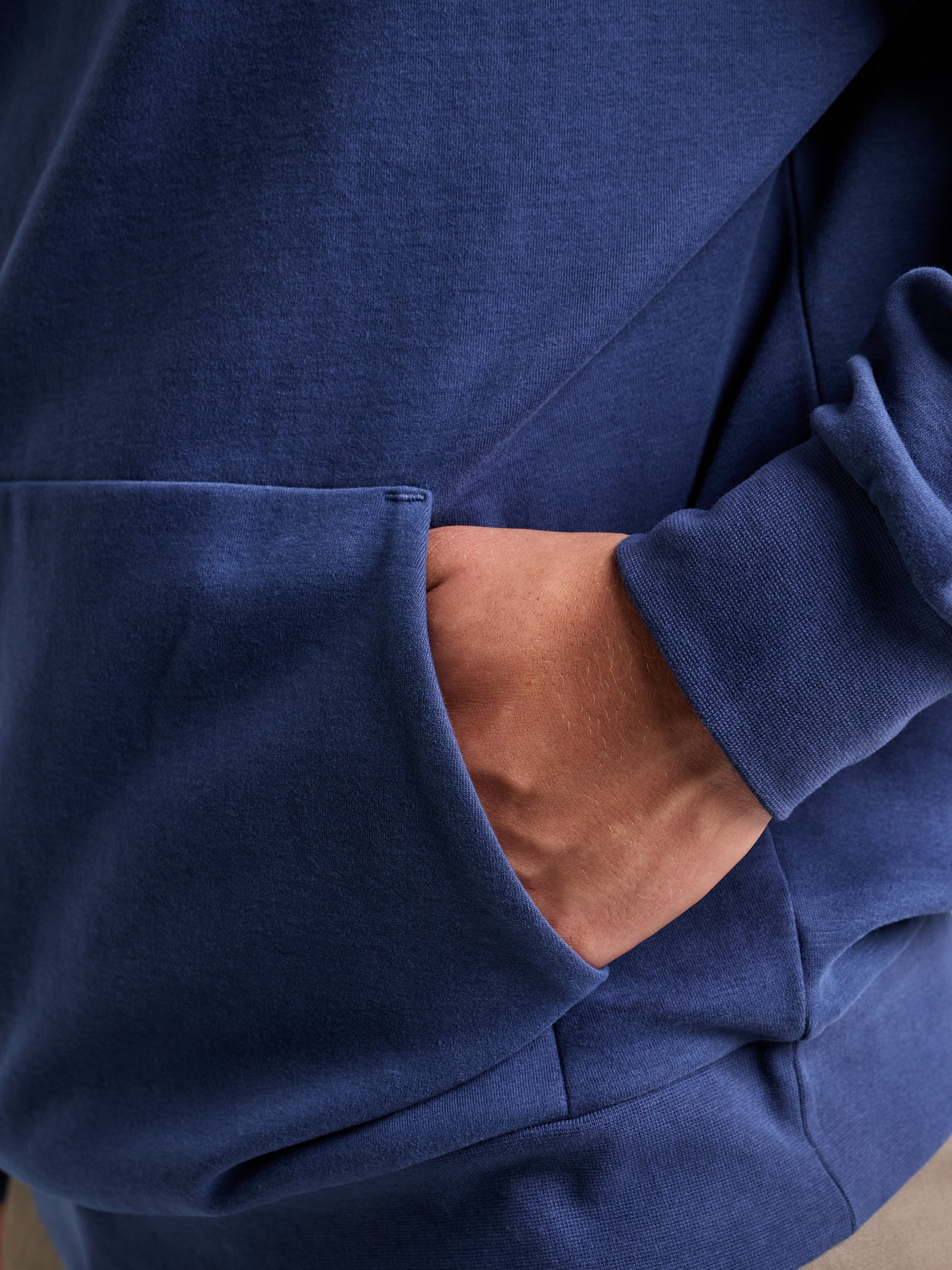 A person wearing a blue SLAM LOGO HOODIE with an adjustable hood, hands tucked into the front pocket. The image is focused on the pocket and hand, with the sleeve slightly rolled up to subtly showcase the SLAM logo.