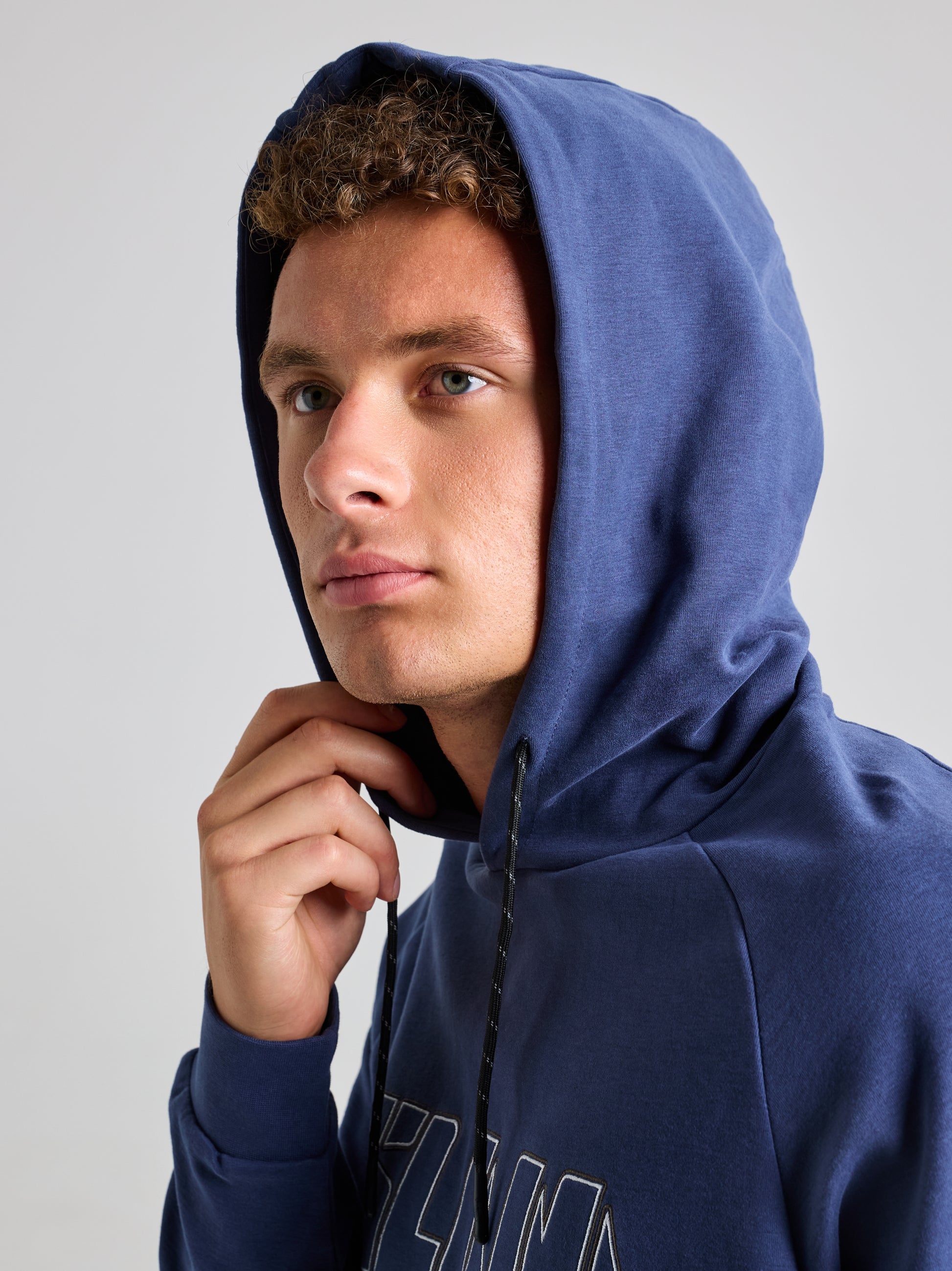 A young person with curly hair is wearing the SLAM LOGO HOODIE, hood up. They are gazing slightly upward with a thoughtful expression against a plain background.