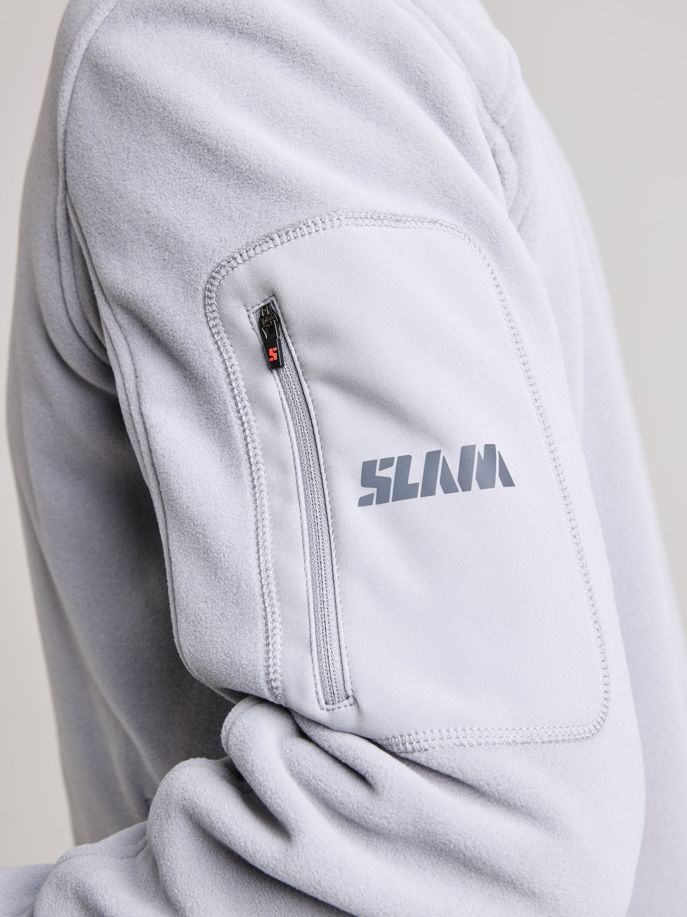 Close-up of a sleeve from the DECK FLEECE jacket in light gray, featuring a small zippered pocket with "SLAM" printed on it, crafted from recycled microfibre.