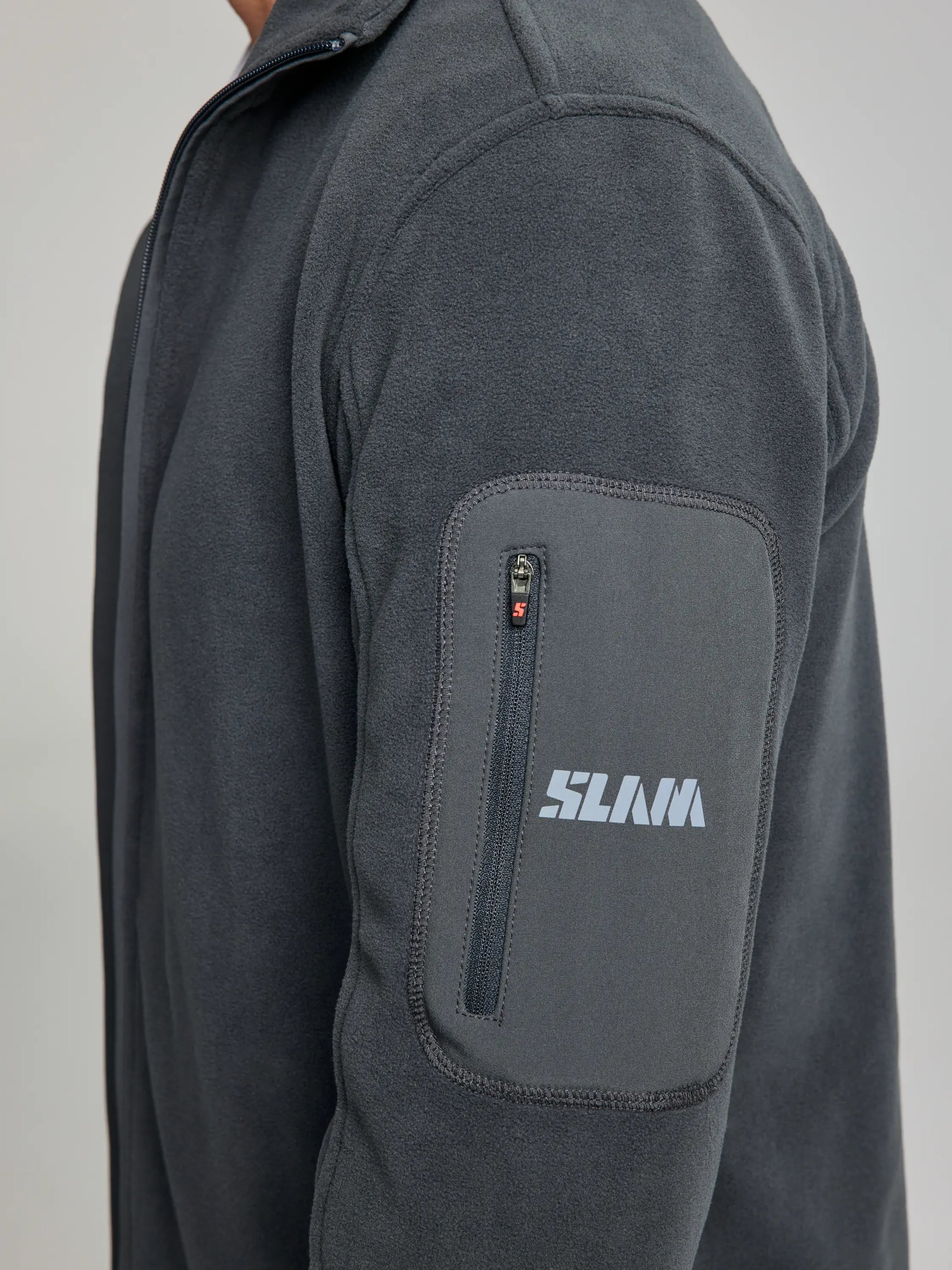 Close-up of a person wearing the dark gray DECK FLEECE CL jacket by SLAM, featuring a convenient zipper pocket on the sleeve with "SLAM" printed in light blue letters, seamlessly combining style and functionality in every detail.