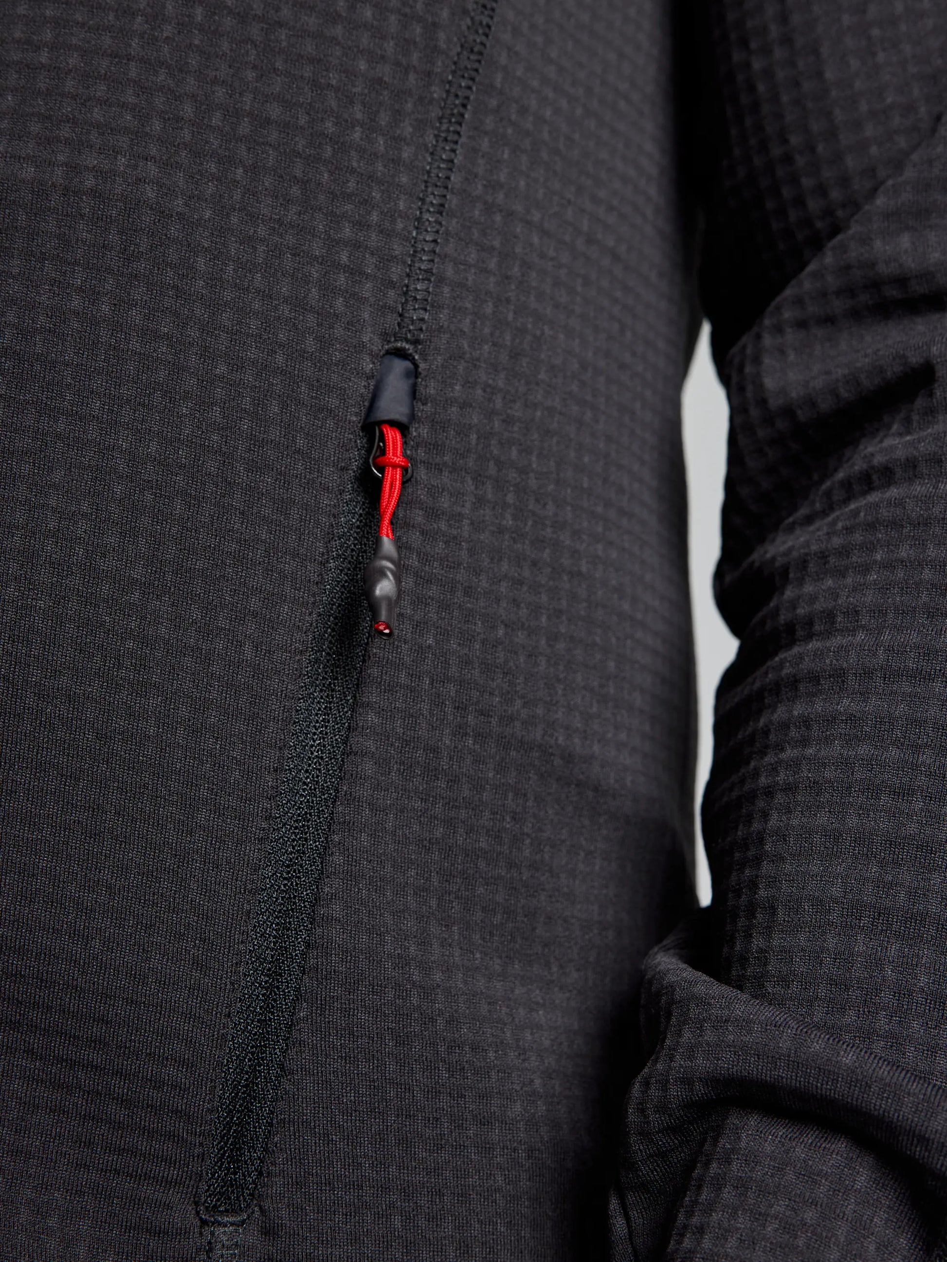 A close-up image showcases the ACTIVE GRID FLEECE by SLAM, highlighting its breathable black textured fabric. A zippered pocket with a primarily black zipper is accented by a red pull, and the Grid Fleece pattern adds dimension. Part of an arm is subtly visible on the right side of the image.