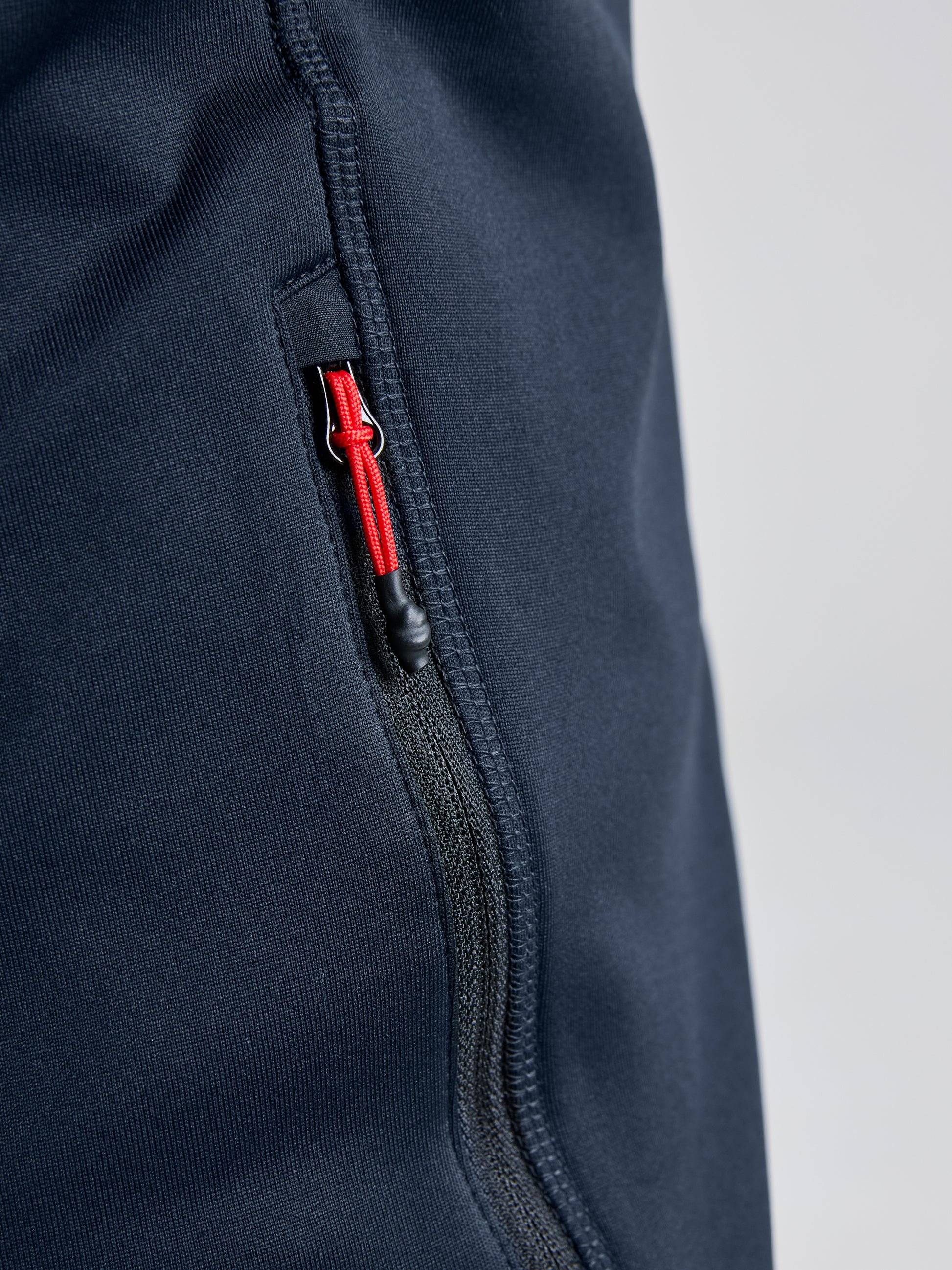 Close-up of a black zipper on navy blue fabric, potentially crafted from recycled materials. The zipper includes a small red pull tab with a black cord, complementing the textured look of SLAM's ACTIVE FLEECE POWERSTRETCH garments, such as jackets or pants, highlighting their elasticity.