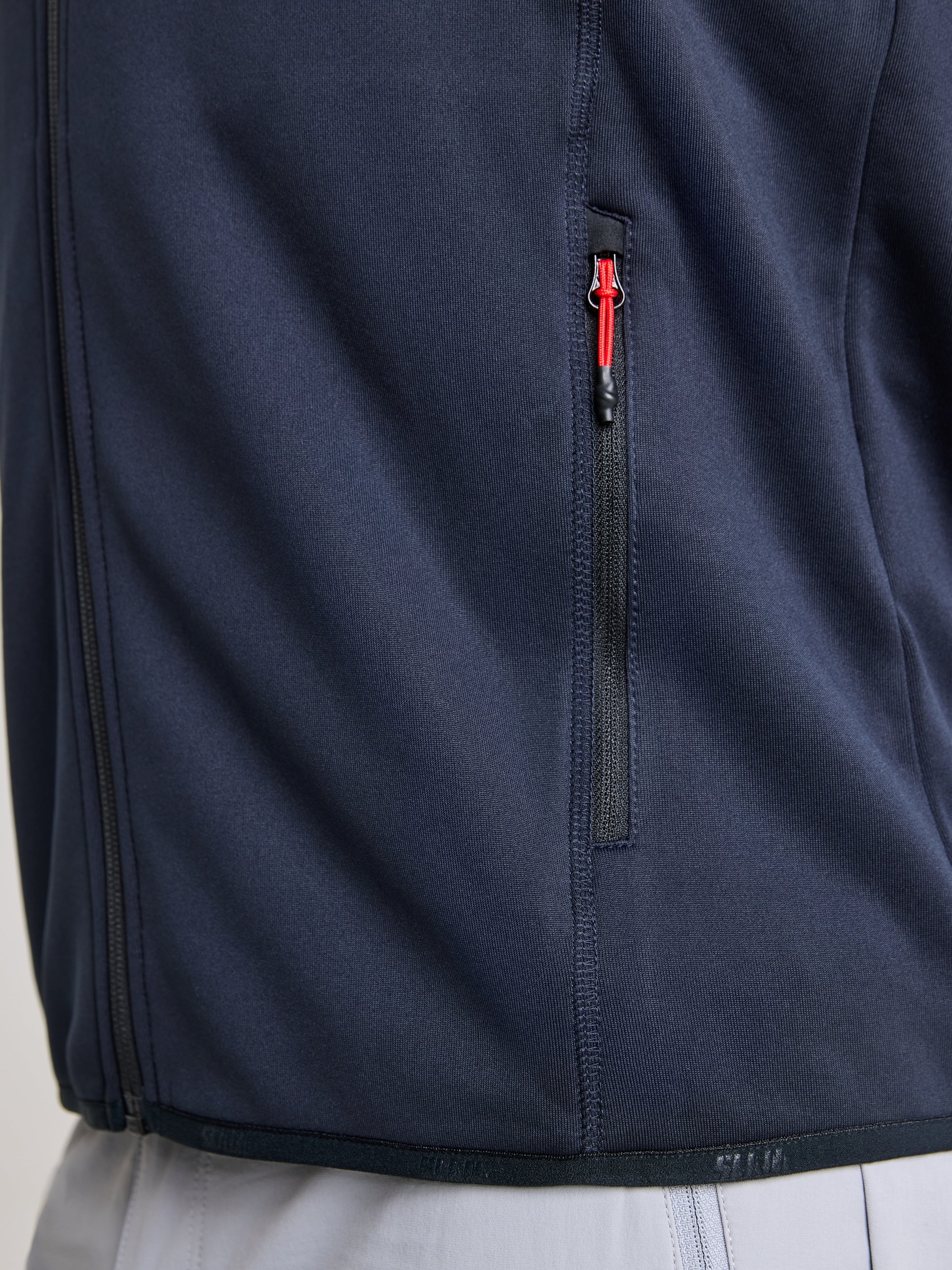 A close-up of a person wearing the SLAM ACTIVE FLEECE POWERSTRETCH jacket in dark blue, crafted from recycled fabric. The jacket features a striking red zipper on the pocket and is paired with light-colored pants. The focus is on the side of the jacket, emphasizing its elasticity and textured design details.