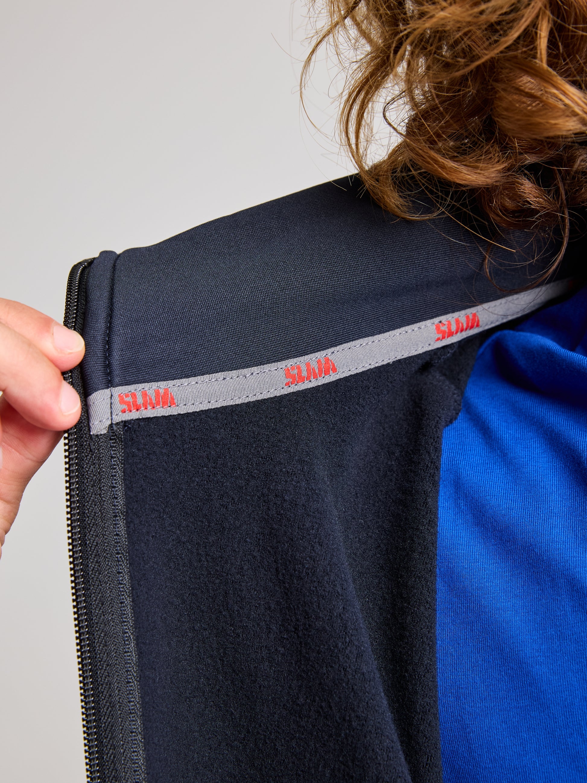 A person wearing the ACTIVE FLEECE POWERSTRETCH jacket by SLAM, with an open collar that reveals a blue shirt beneath. The inner collar features a gray strip with "SLAM" in red text. An elastic hand grips the collar, highlighting the jacket's enhanced comfort and flexibility.