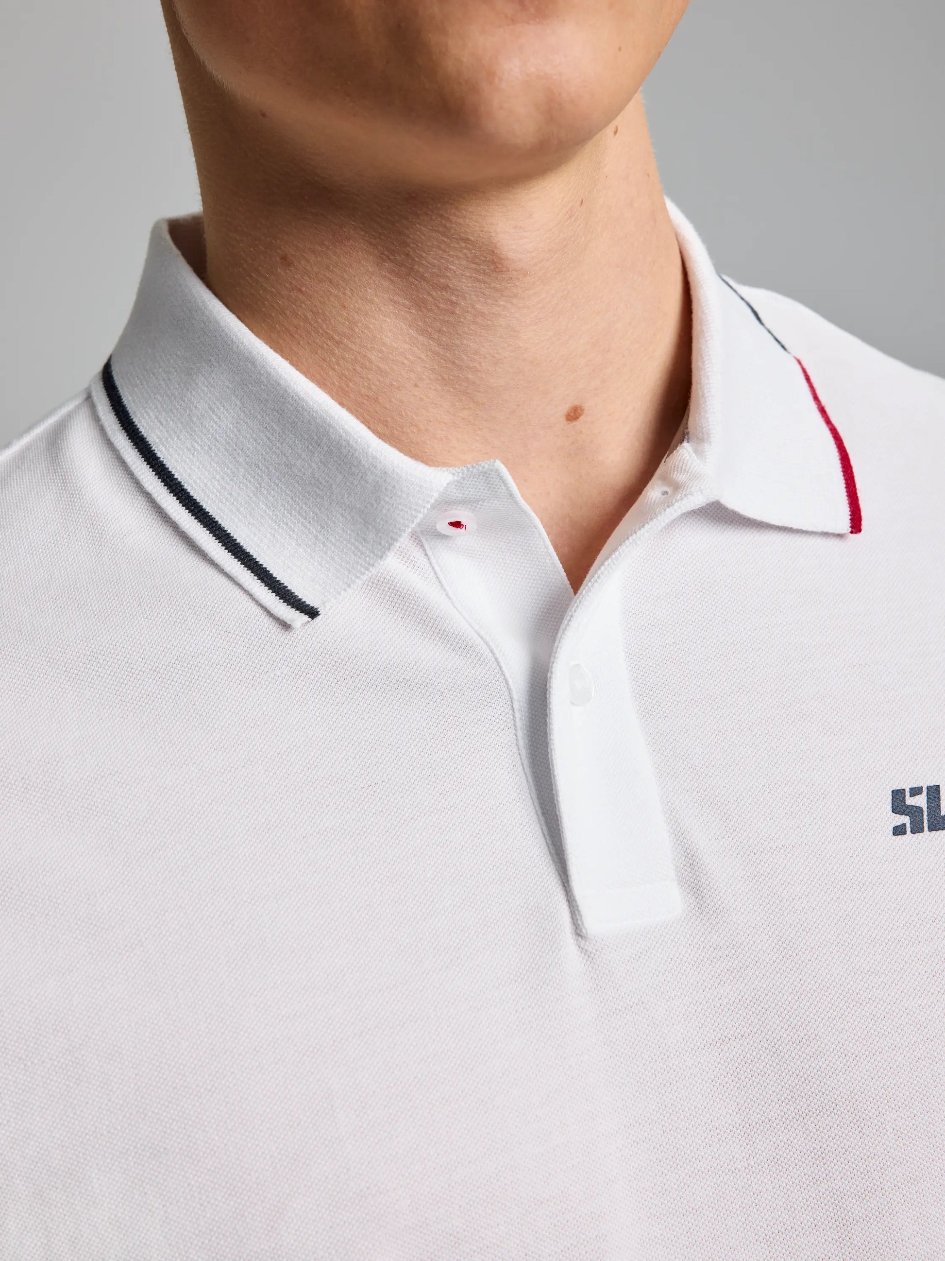 A close-up showcases a person wearing the SLAM POLO AEGIR SHORT SLEEVE CL in white, featuring a small embroidered logo on the chest. The ribbed collar is accented with thin black and red stripes, giving it a nautical flair, and the shirt is fully buttoned up. The background is set against a neutral gray tone.