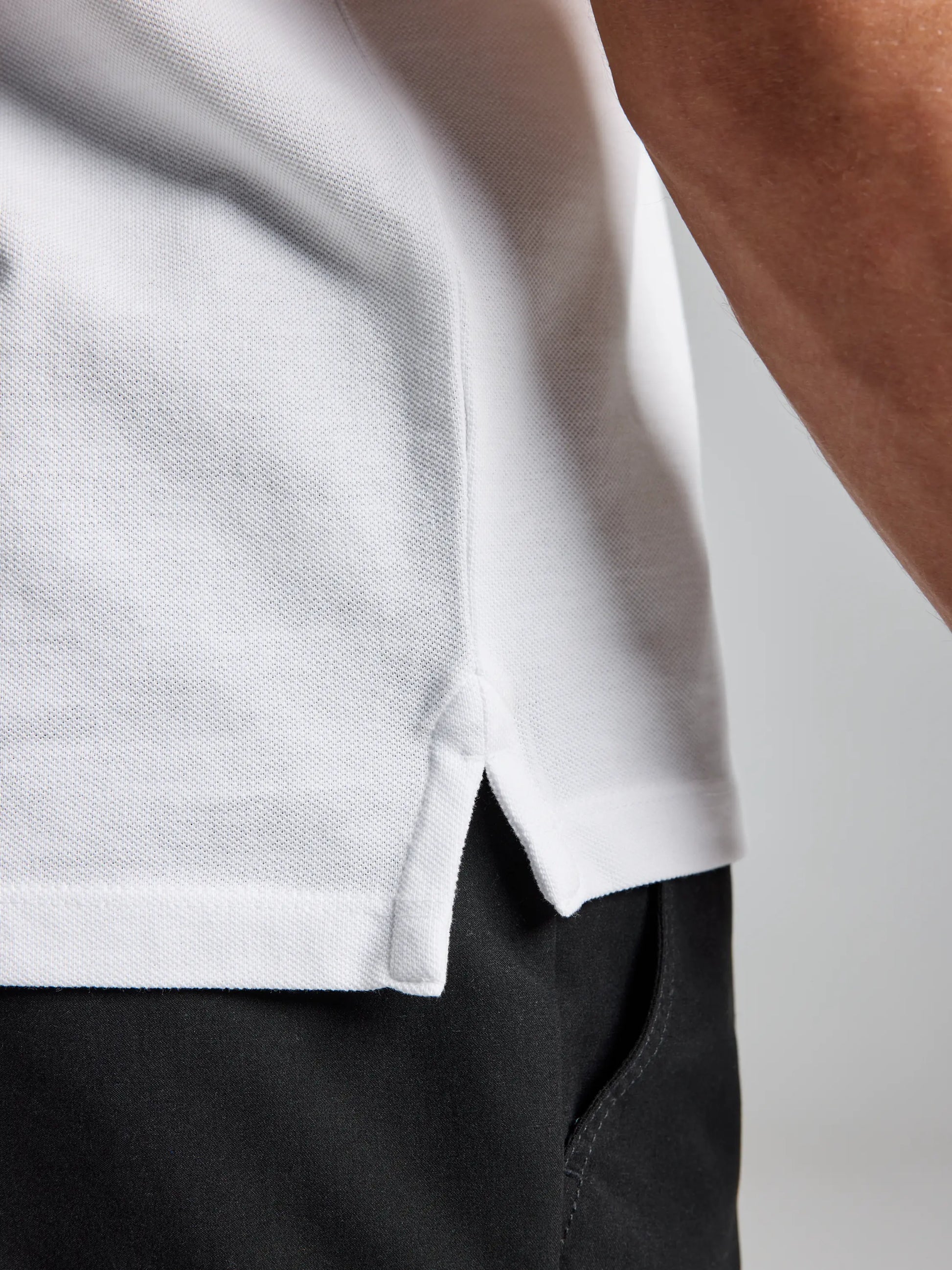 A close-up of an individual wearing the SLAM POLO AEGIR SHORT SLEEVE CL, which features a ribbed collar and is paired with black pants. The side seam and hem of the white polo shirt reveal a small notch at the hemline, with the fabric's texture subtly visible, enhancing its modern nautical style appeal.