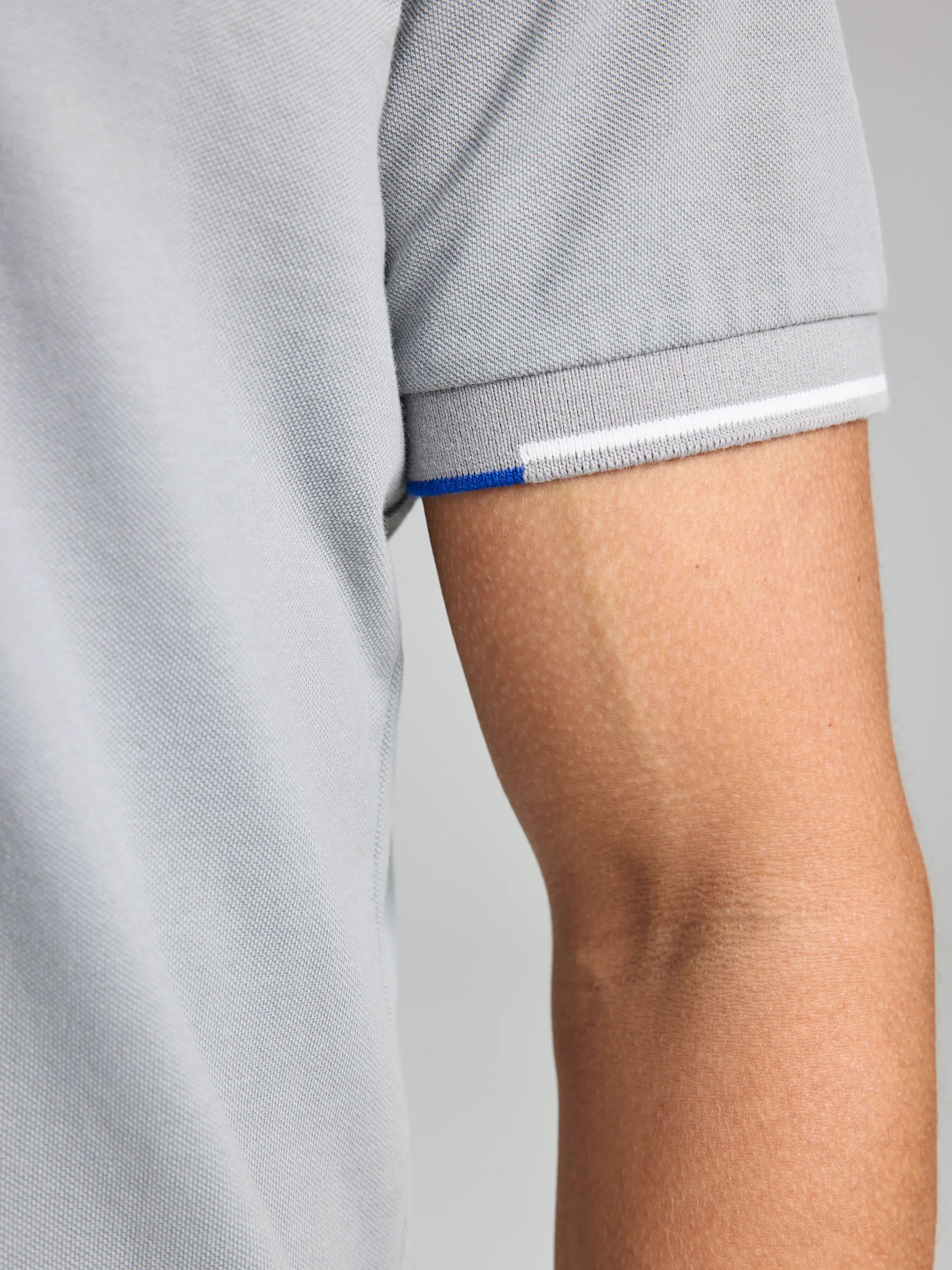 Close-up of a person's arm dressed in the SLAM POLO AEGIR SHORT SLEEVE CL, featuring a light gray color. The sleeve showcases a subtle blue and white stripe at the edge, hinting at nautical style, against a plain, soft-focused background that enhances the look.