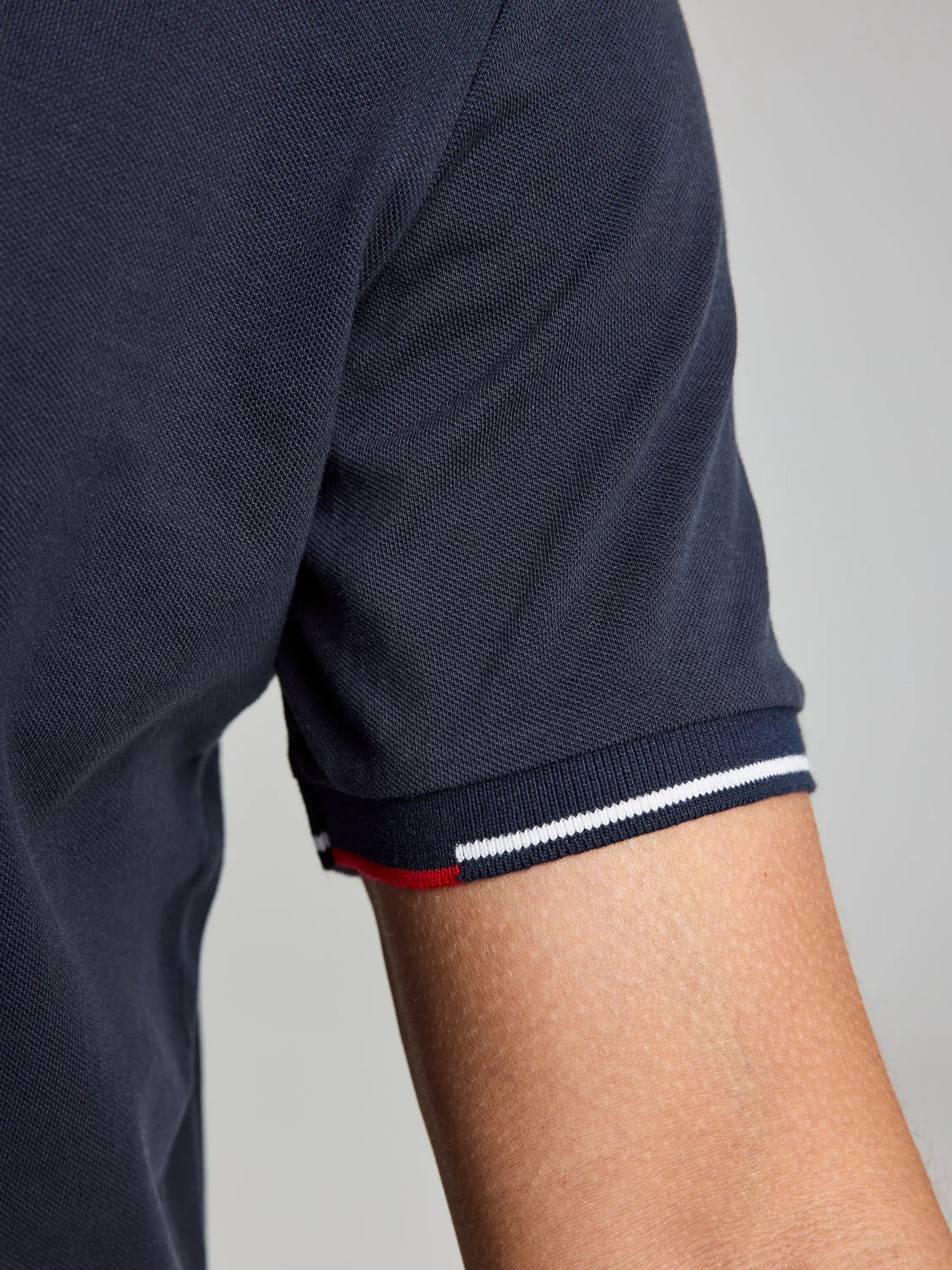 A close-up of a person's arm wearing the SLAM POLO AEGIR SHORT SLEEVE CL in nautical-style dark blue. The ribbed collar features red, white, and blue striped trim against a neutral gray background.