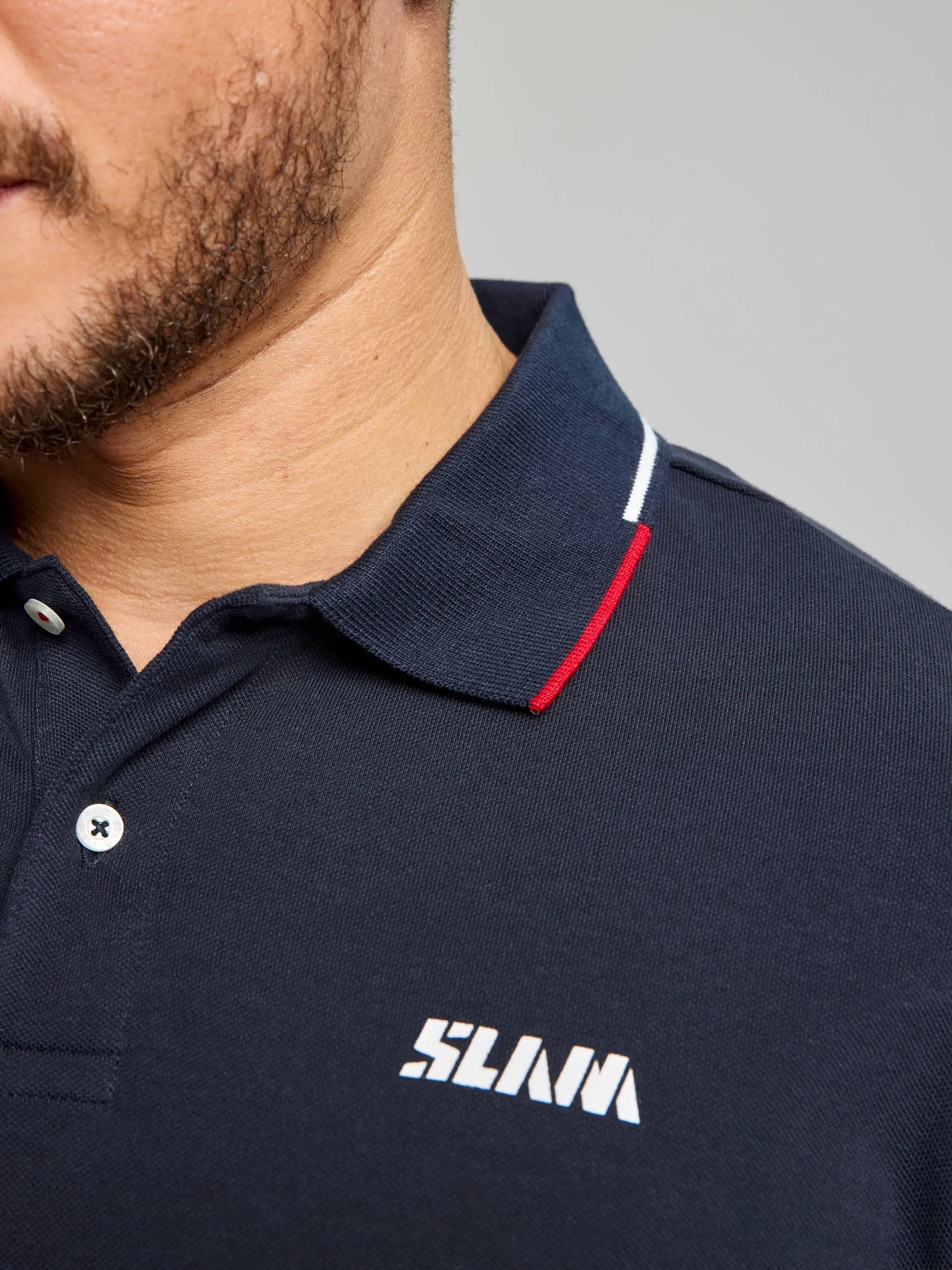 Close-up of a person in the POLO AEGIR SHORT SLEEVE CL by SLAM, featuring a nautical-style design. The ribbed collar showcases red and white stripes, complementing the brand logo on the chest. A stubbled chin is partially visible.