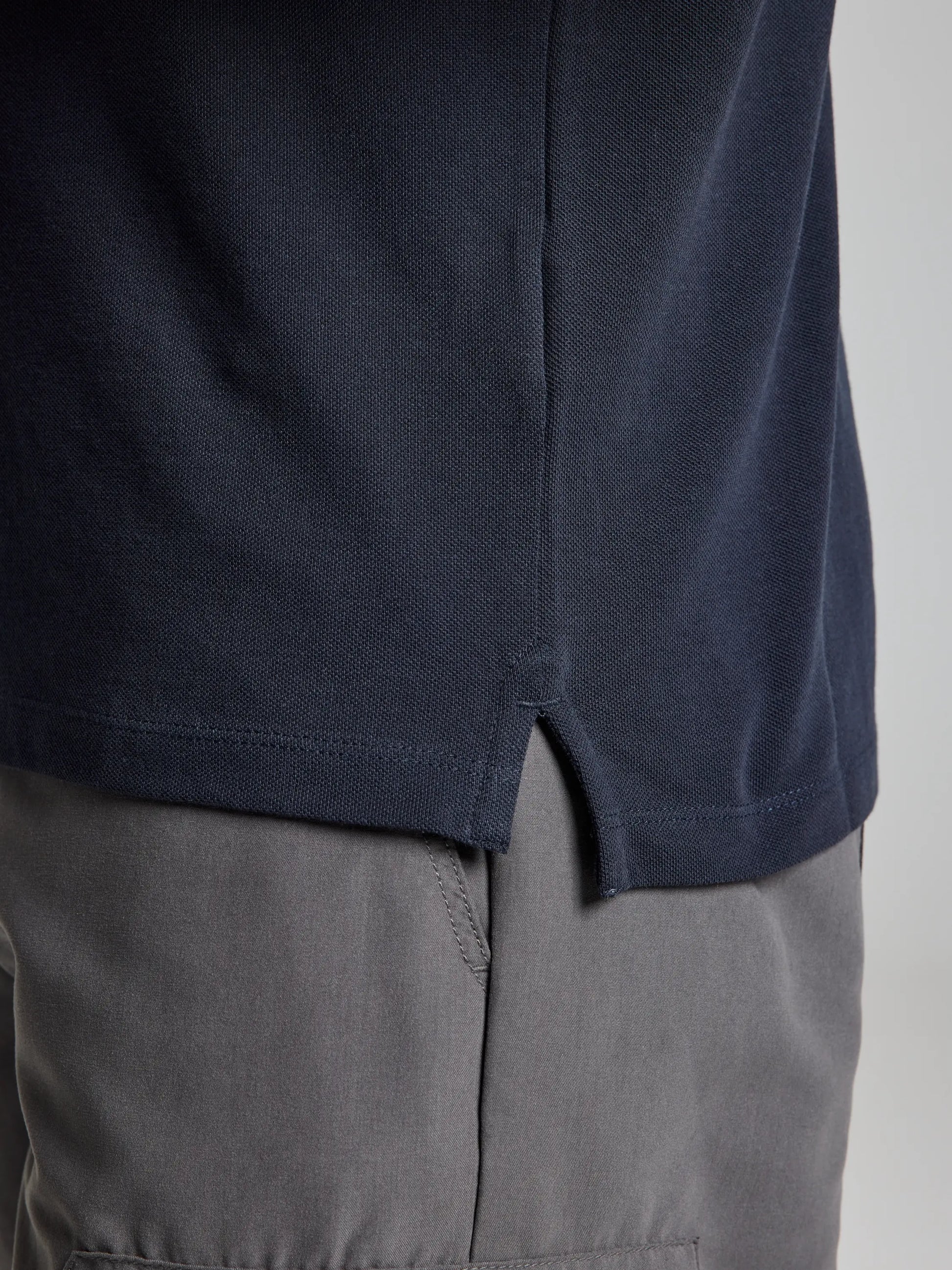 Close-up view of a person wearing the POLO AEGIR SHORT SLEEVE CL by SLAM, featuring a dark blue color with a ribbed collar and side slit, paired with gray pants. The image focuses on the midsection, highlighting the textiles and details of the clothing.