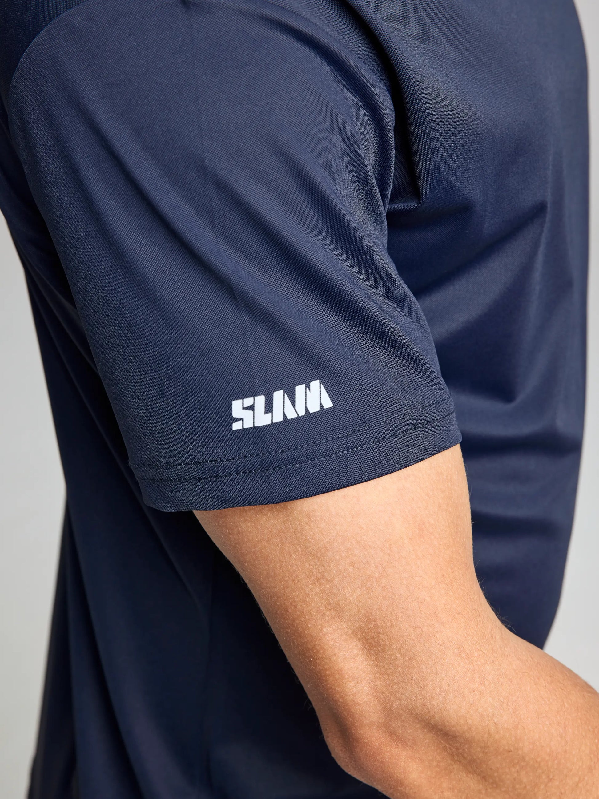 A person wearing a dark blue short-sleeve shirt from the SLAM brand, known as the TECH PIQUE T SHIRT CL, crafted from breathable stretch techno pique fabric. The word "SLAMA" is prominently printed in white on the sleeve. The focus is on the shirt's UV-resistant feature covering the upper arm and part of the side view.