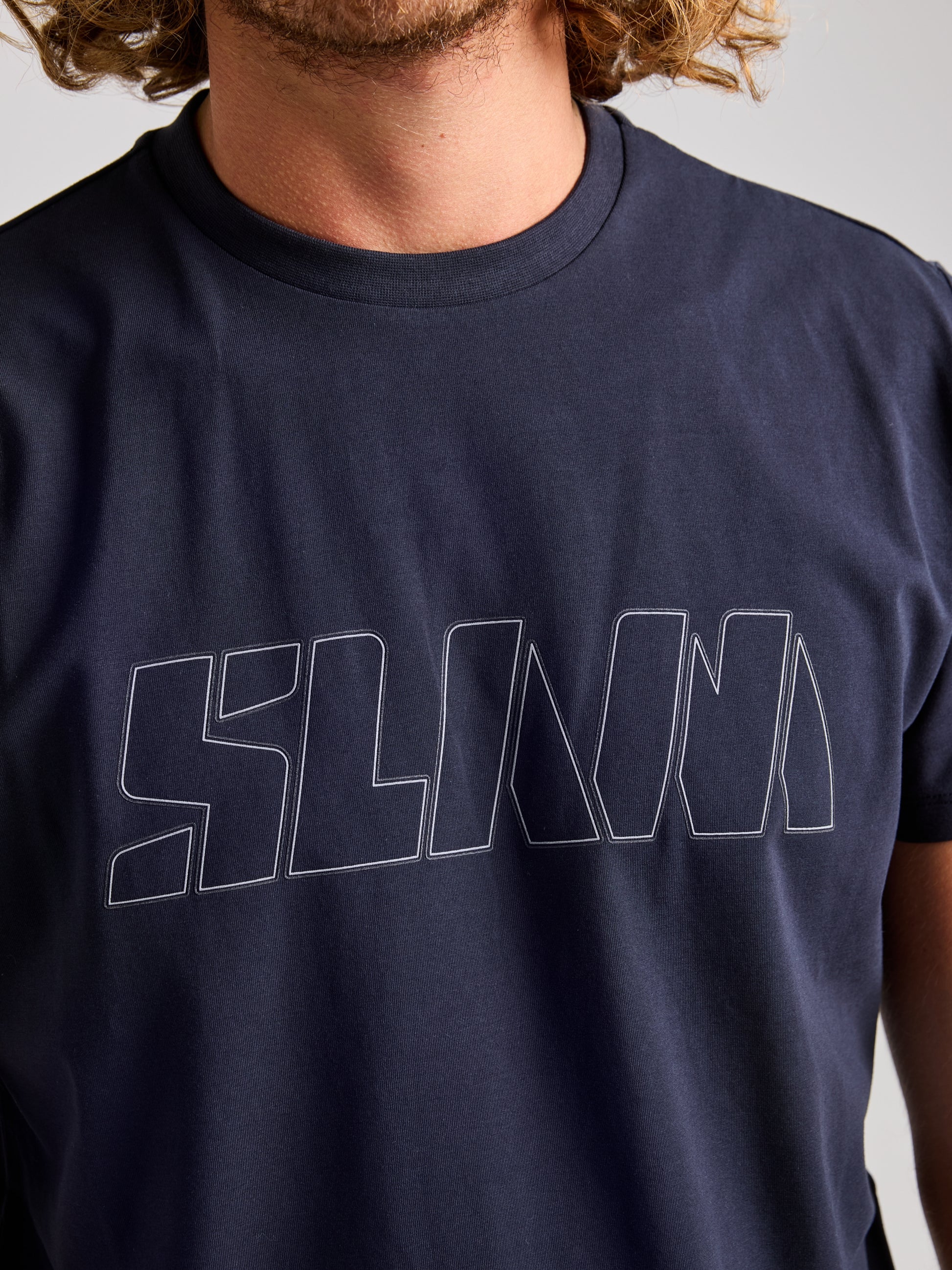 A person with curly hair is wearing the SLAM brand navy blue LOGO T SHIRT, which features reflective "SLAM" lettering in large, outlined uppercase on the chest. This standout piece is made from stretch jersey cotton and contrasts sharply against a simple, light-colored background.