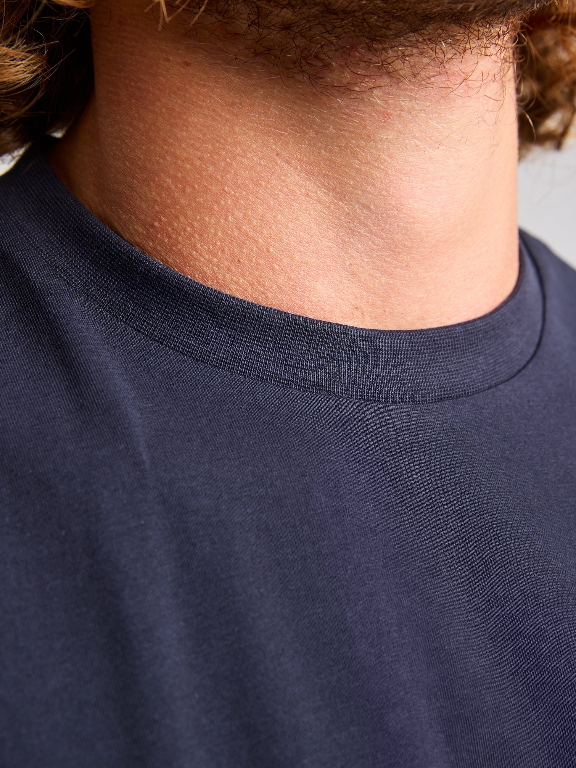 A close-up shot features a person wearing SLAM's dark blue LOGO T SHIRT, emphasizing the neckline and part of the neck. Made from stretch jersey cotton, the fabric looks soft and high-quality. The top of the image shows a hint of hair, highlighting its modern reflective print.