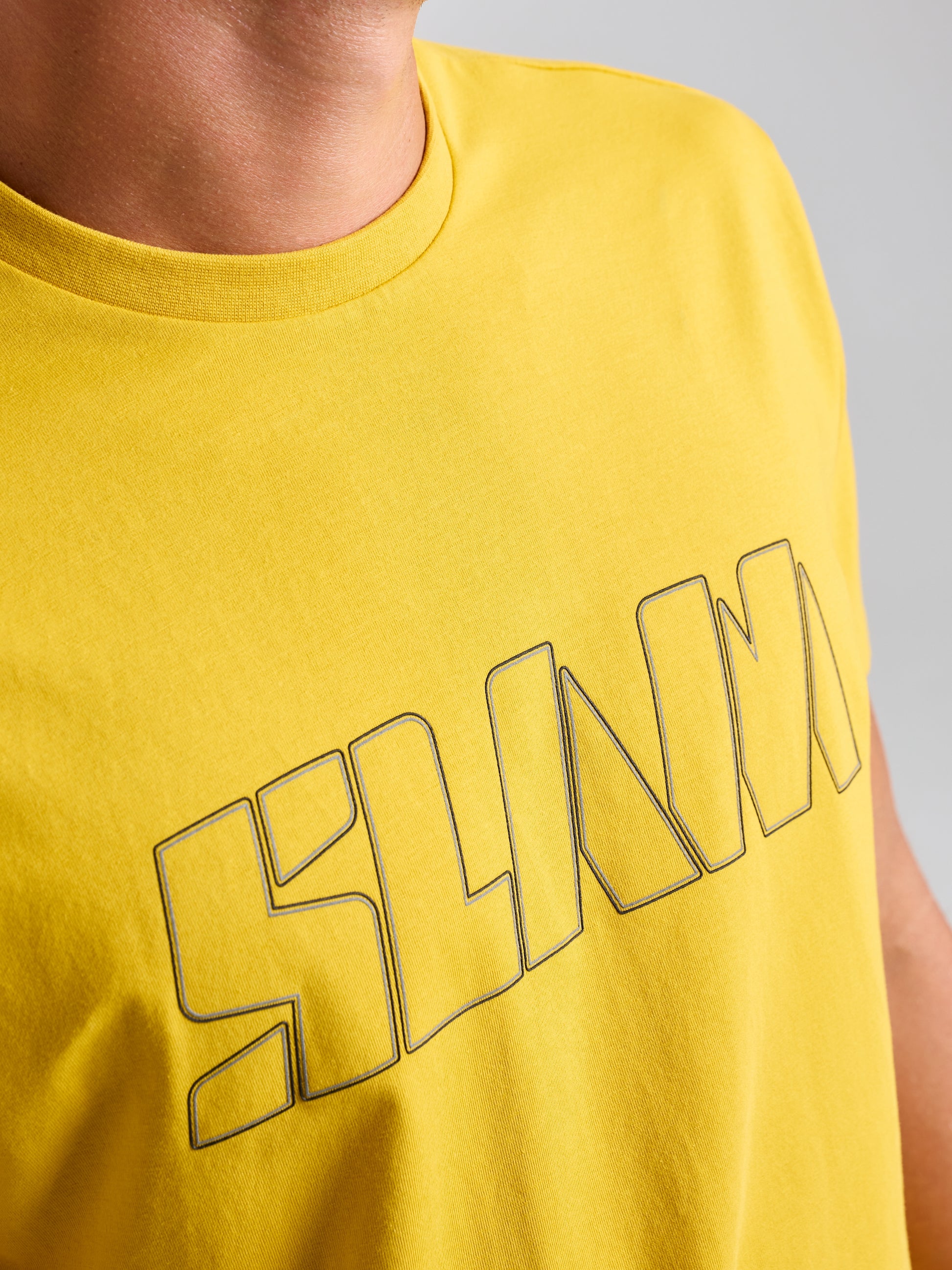 A close-up image features a person wearing the LOGO T SHIRT by SLAM, a bright yellow men's tee made from stretch jersey cotton. The word "SLAM" appears prominently in large, outlined letters across the chest against a neutral gray background.