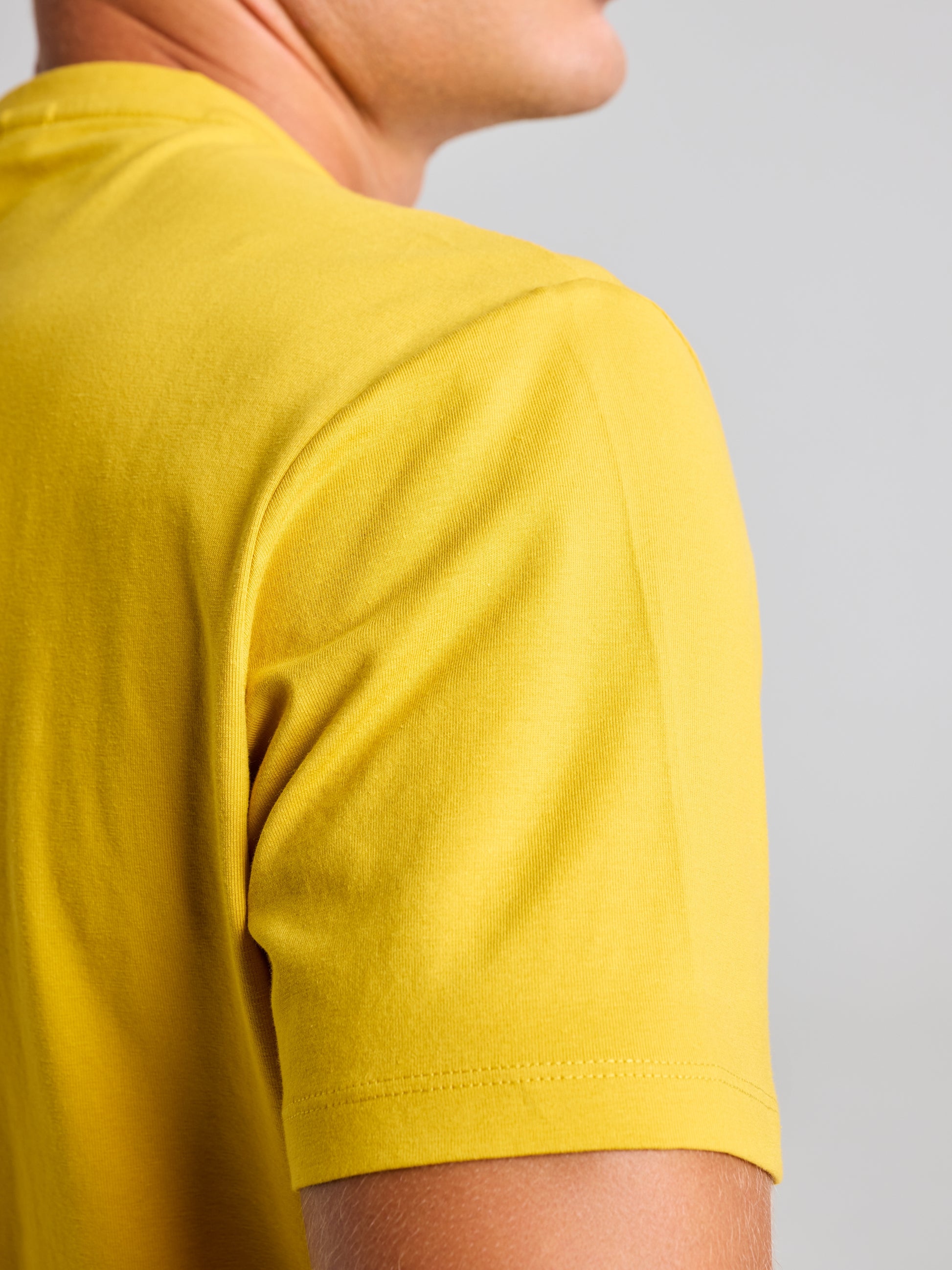 A person wearing a yellow LOGO T SHIRT by SLAM, with emphasis on the upper arm and shoulder. The smooth stretch jersey cotton fabric contours perfectly to the skin, highlighting a sleek silhouette against a plain, neutral background.