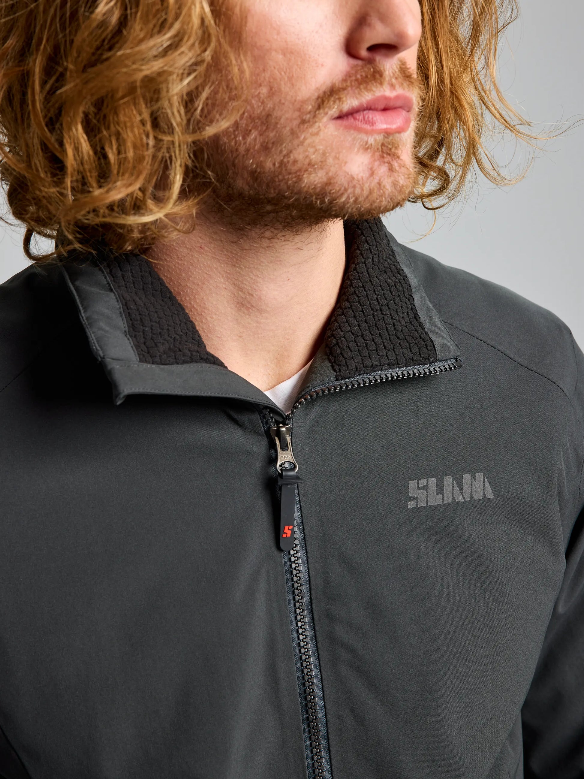 An individual with shoulder-length wavy hair is sporting a dark gray, waterproof DOCK INSULATED JACKET CL from SLAM, which comes with a fleece-lined collar. The men's nautical winter jacket showcases a small brand logo on the left side. The person's face is partially visible as they look slightly to the side.