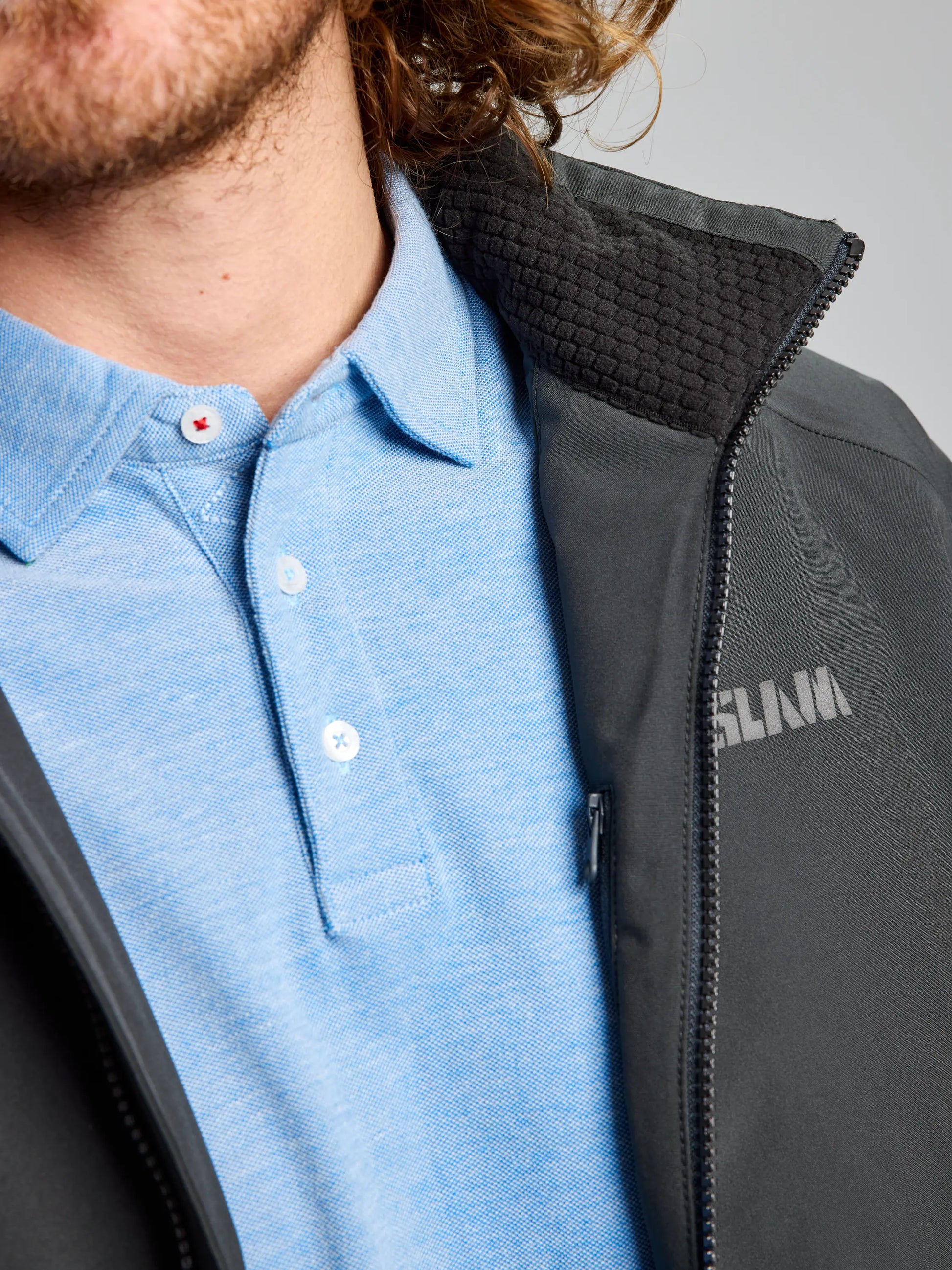 A close-up image shows a person wearing a light blue collared shirt beneath the SLAM DOCK INSULATED JACKET CL, with "SLAM" visible. The fleece-lined collar emerges slightly, as curly hair partially obscures their face. The waterproof texture and zipper of the jacket are in clear focus.