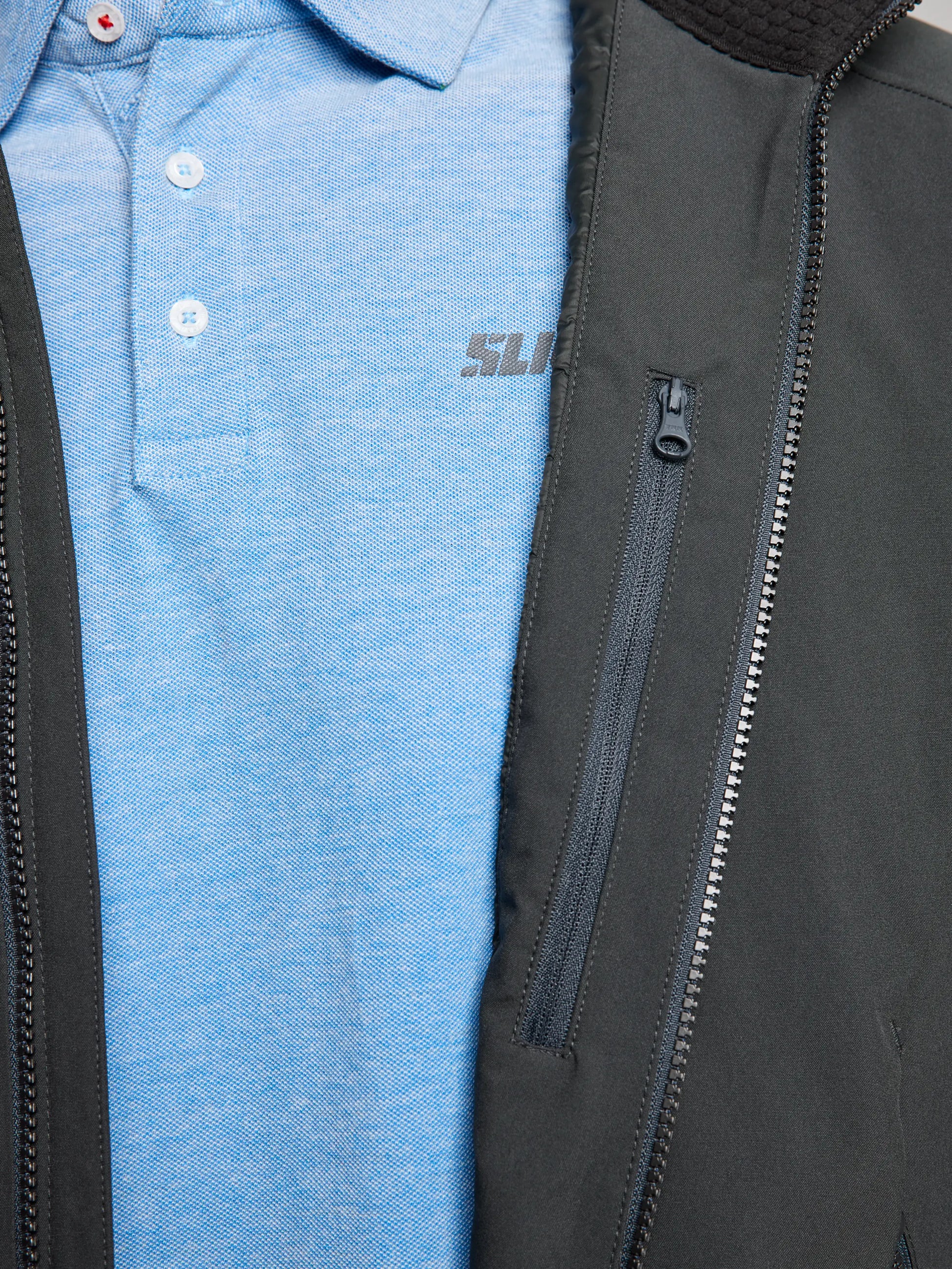 A close-up of a person wearing the DOCK INSULATED JACKET CL by SLAM, featuring a partially zipped black exterior that reveals a light blue polo shirt underneath. The jacket's fleece-lined collar and chest zipper pocket enhance both its stylish appearance and waterproof functionality.