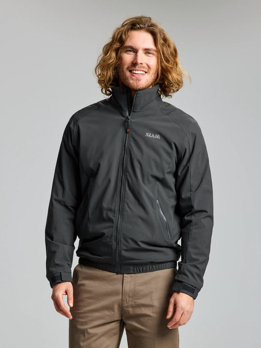 A person with long curly hair is wearing the SLAM DOCK Insulated Jacket CL in graphite and khaki pants, standing against a plain background. The waterproof jacket features a logo on the left side and has a cozy fleece-lined collar.