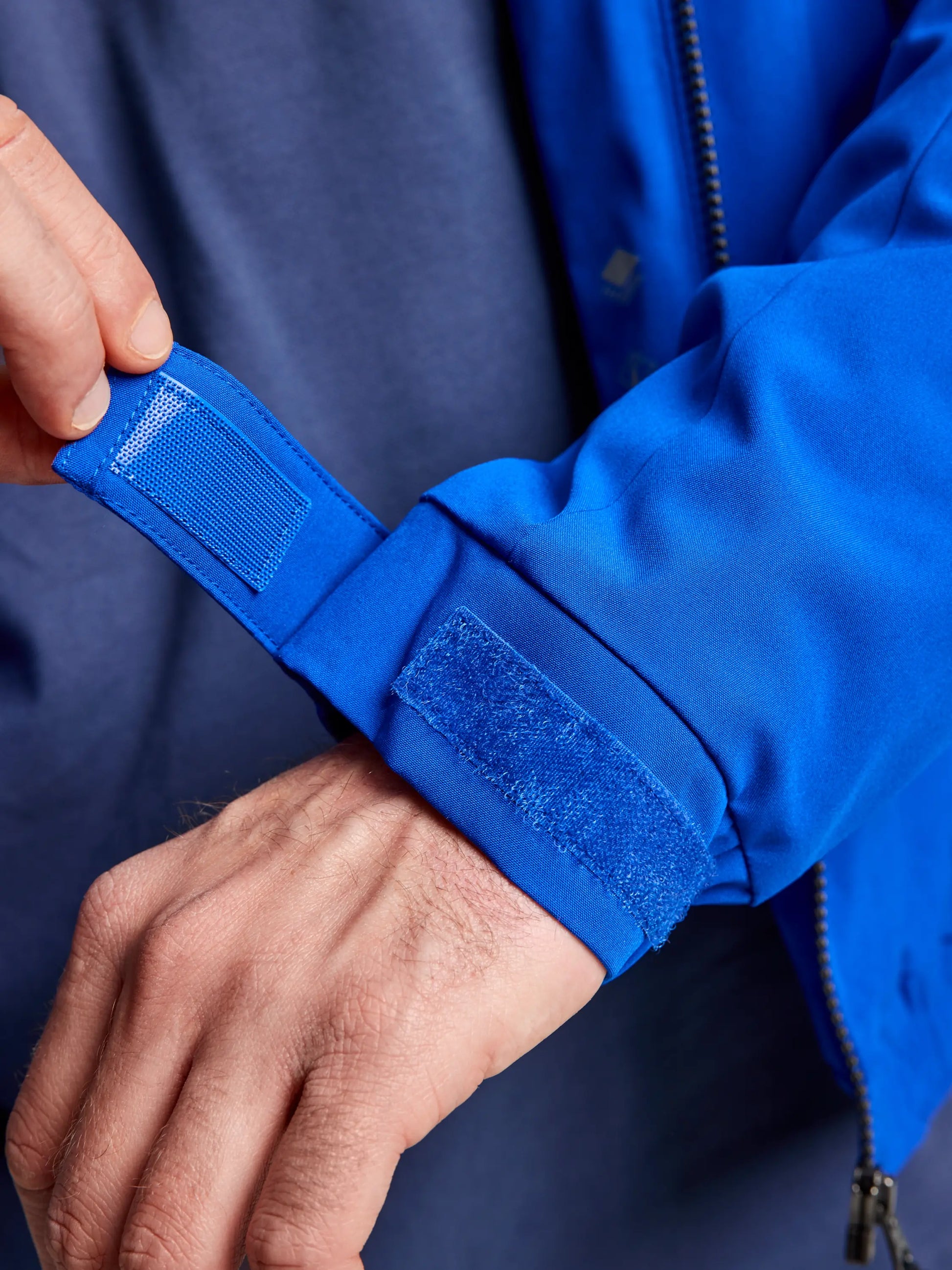 A person adjusts the Velcro strap on the cuff of a blue DOCK INSULATED JACKET CL sleeve by SLAM. The jacket features a zipper and a fleece-lined collar, worn over a blue shirt. The close-up angle highlights the functionality of the adjustable cuff.